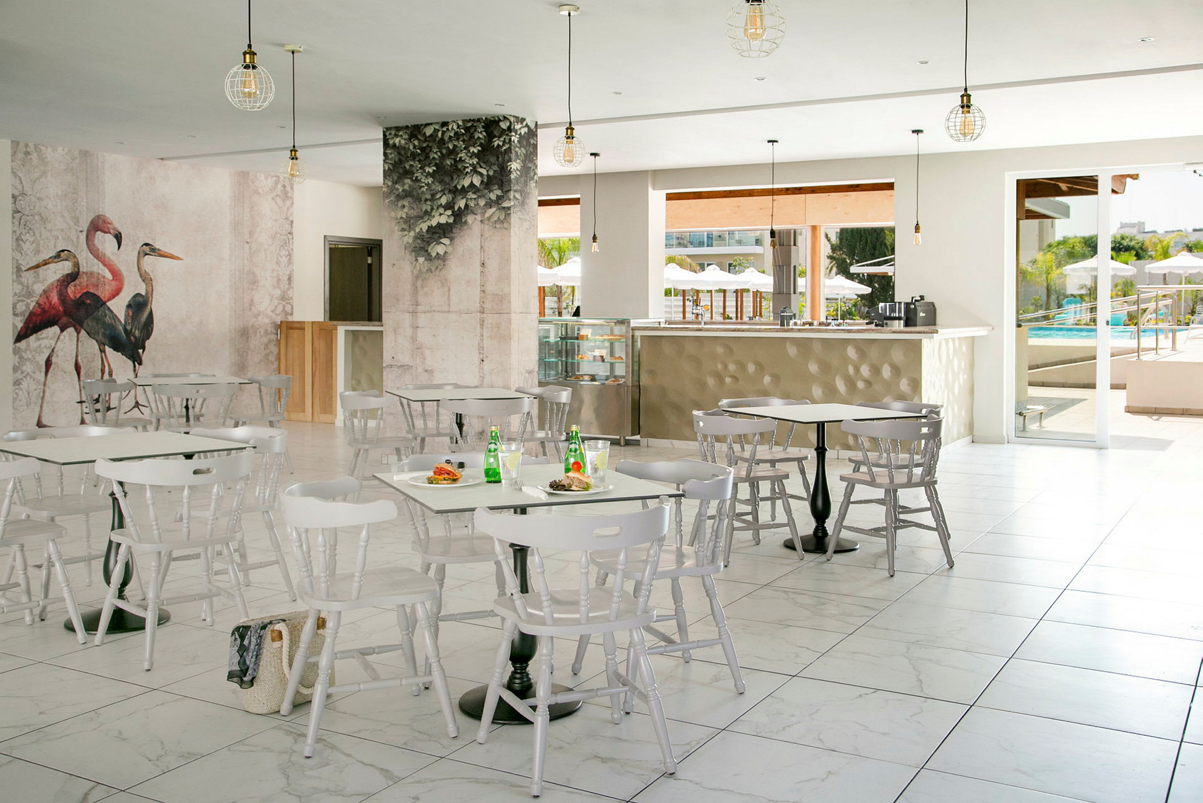 En lys og luftig café med hvide møbler og vægdekorationer med fuglemotiver. I baggrunden er et åbent område, der fører til en pool. (AI-genereret tekst)