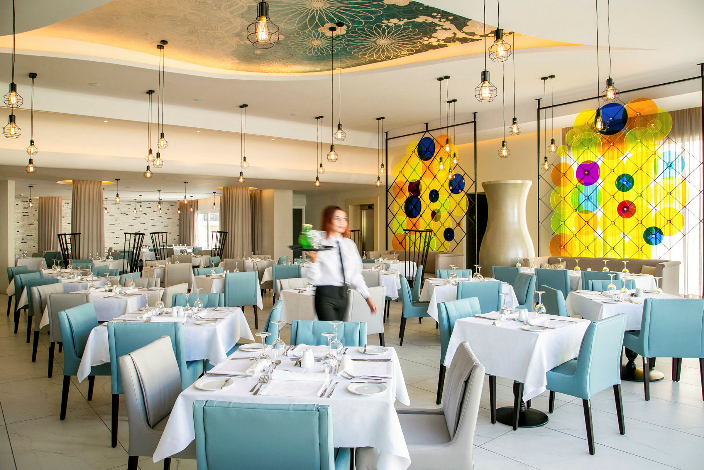 Restaurant med dækkede borde og blå stole samt farverig glasdekoration på væggen. (AI-genereret tekst)