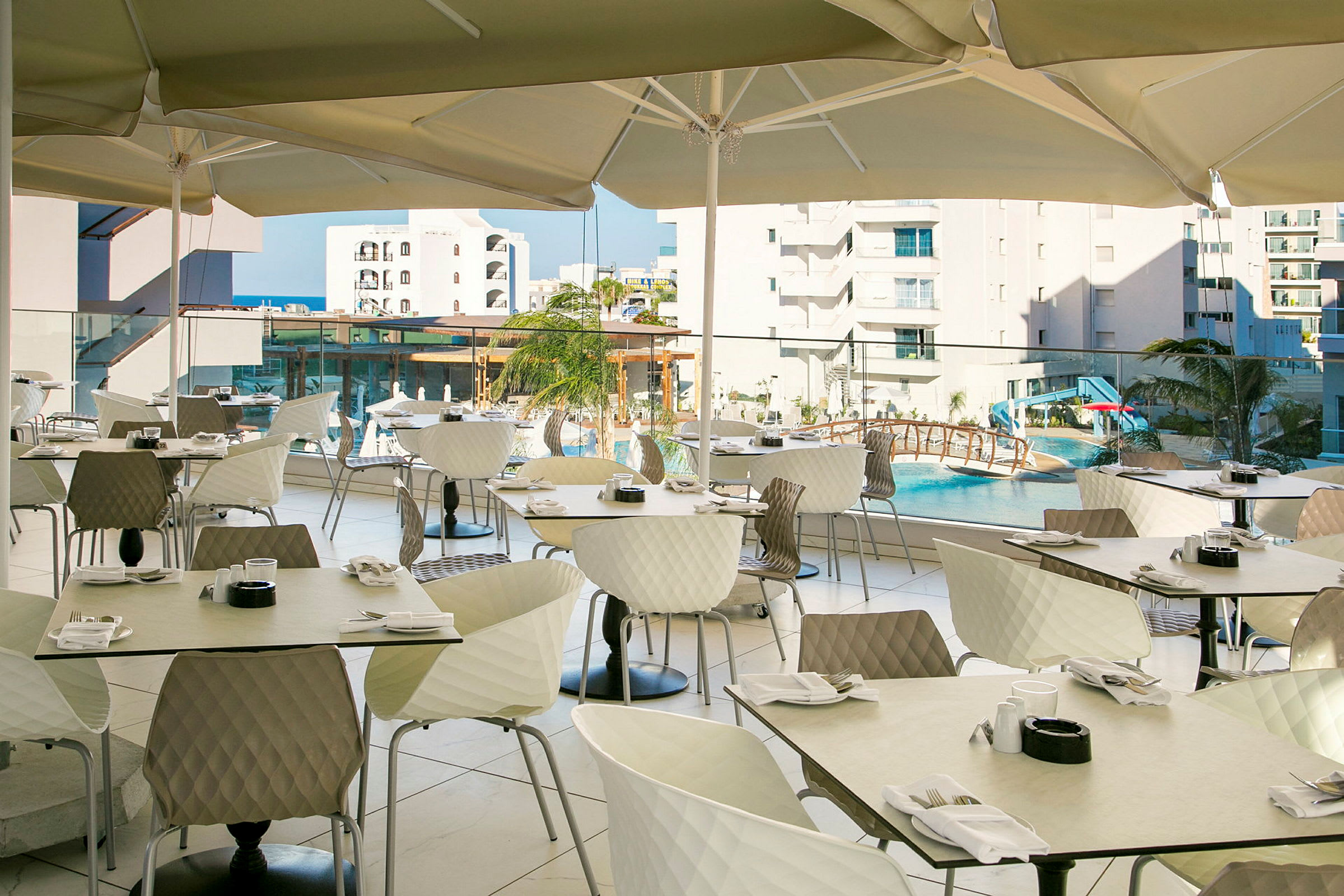 Udendørs restaurant med borde og stole under parasoller, med udsigt over en pool og omkringliggende bygninger. (AI-genereret tekst)