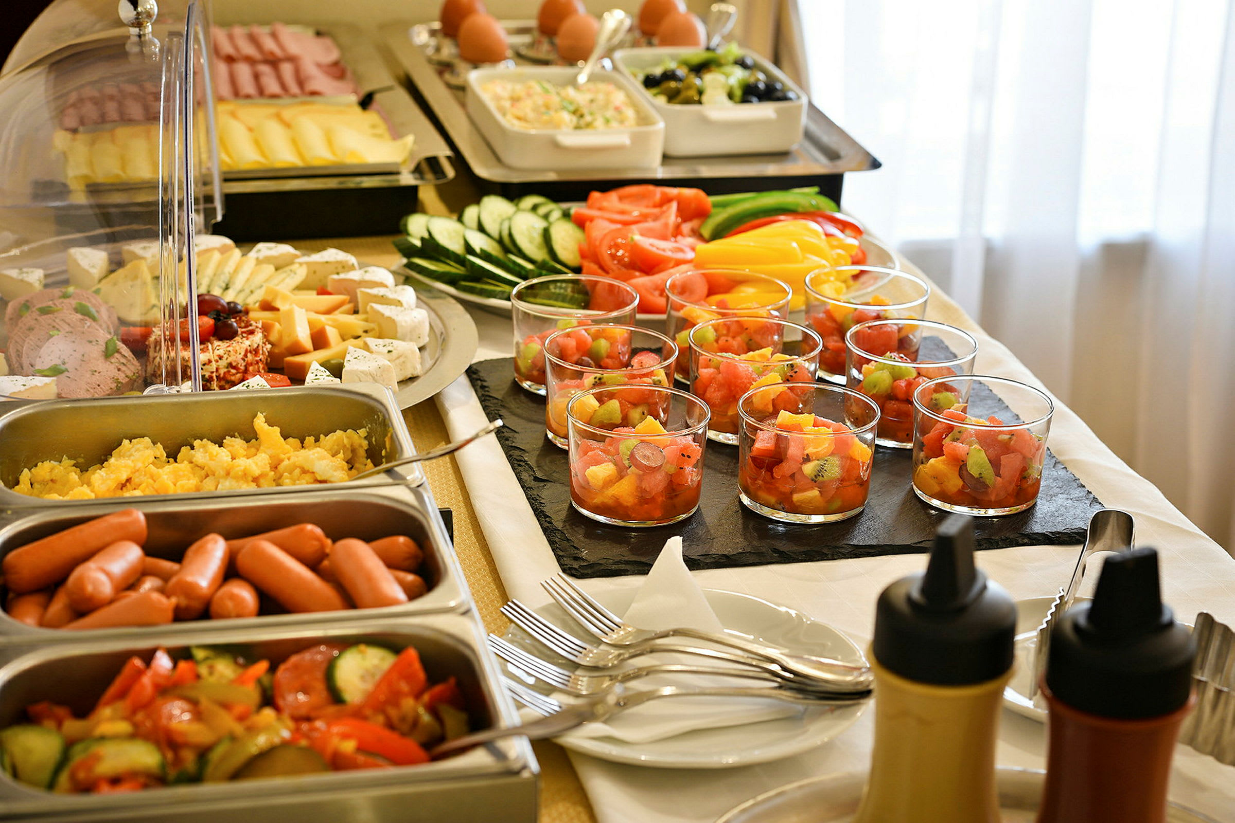 En buffet med forskellige madvarer som pølser, grøntsager, ost, frugtsalat i glas og saucer. (AI-genereret tekst)