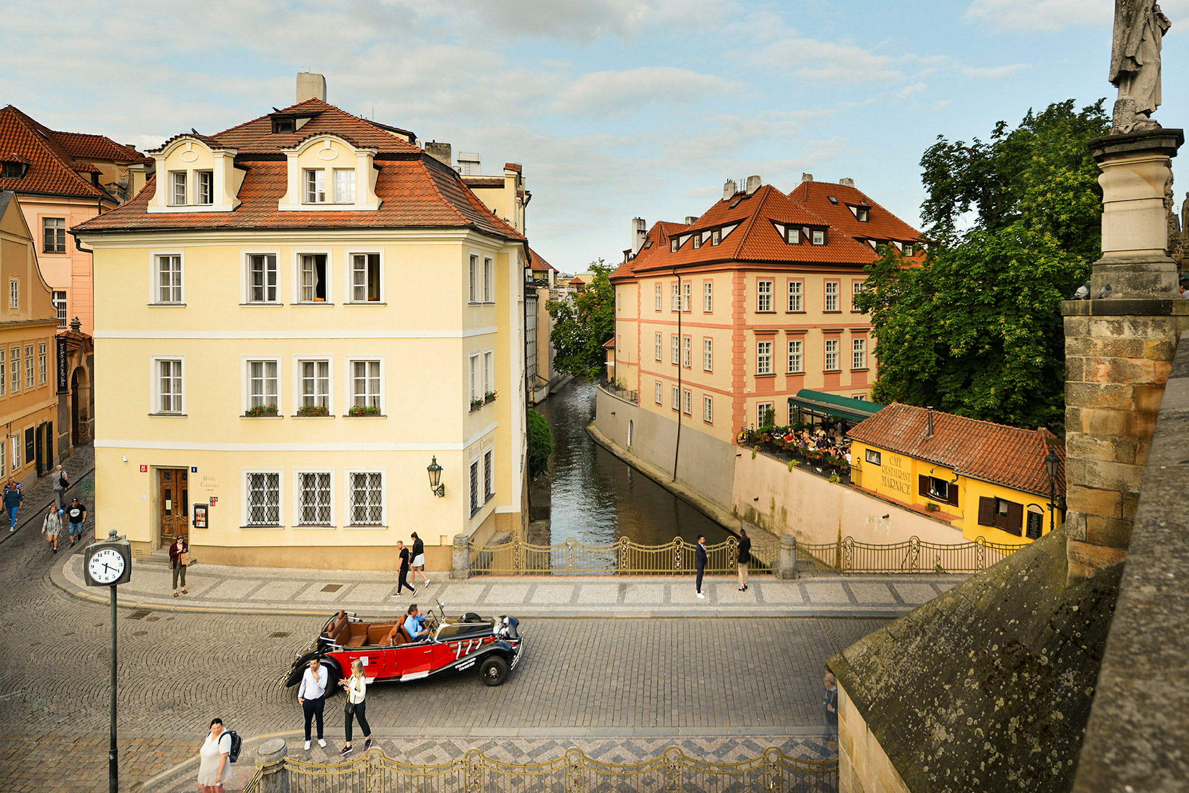 En brostensbelagt gade omgivet af klassiske bygninger med farverige facader. En kanal ses mellem bygningerne samt personer, der går, og en rød bil parkeret på gaden. (AI-genereret tekst)