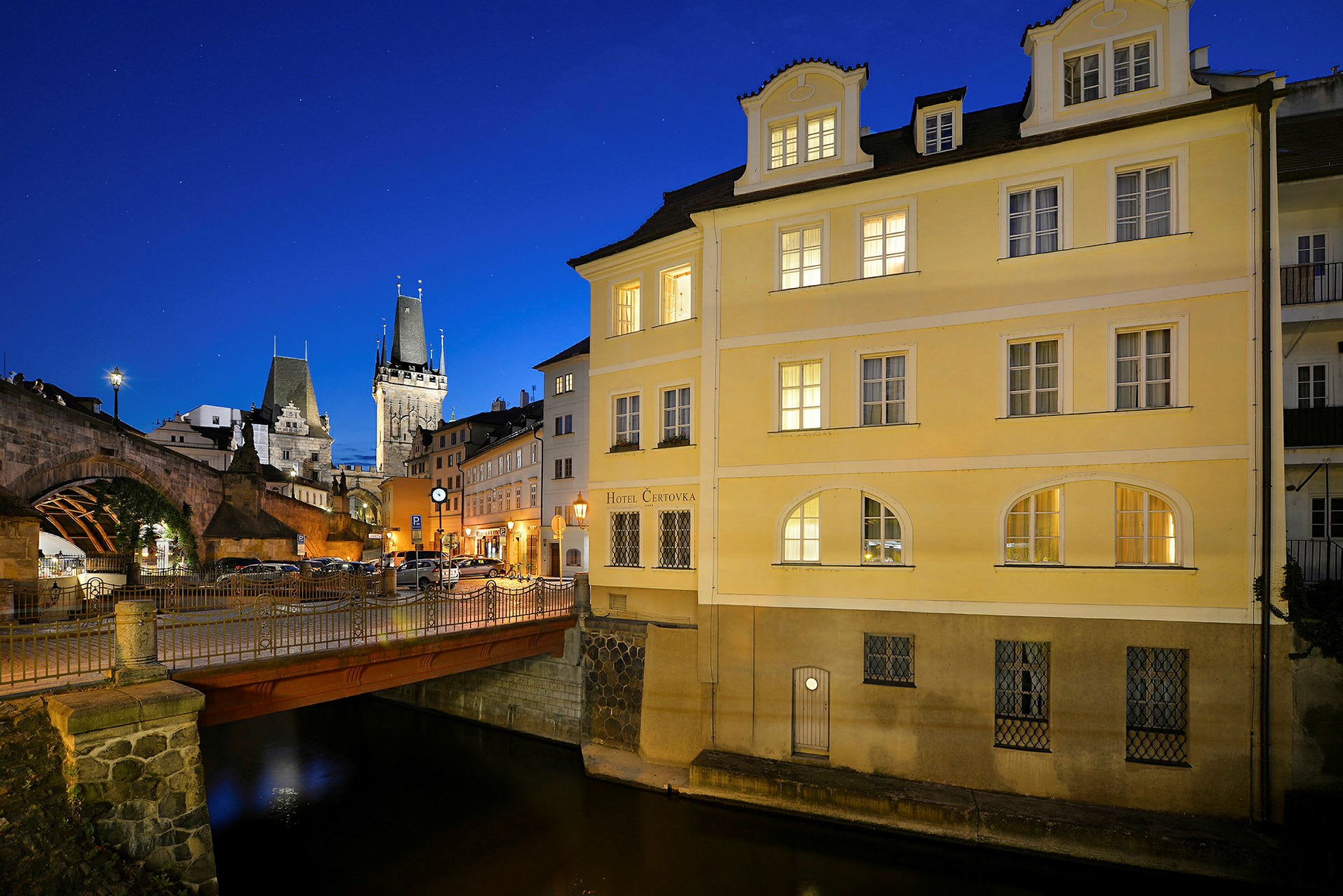 En lille bro over en kanal ved siden af en gul bygning under en blå nattehimmel. I baggrunden ses en historisk stenbro og en kirketårnlignende bygning. (AI-genereret tekst)