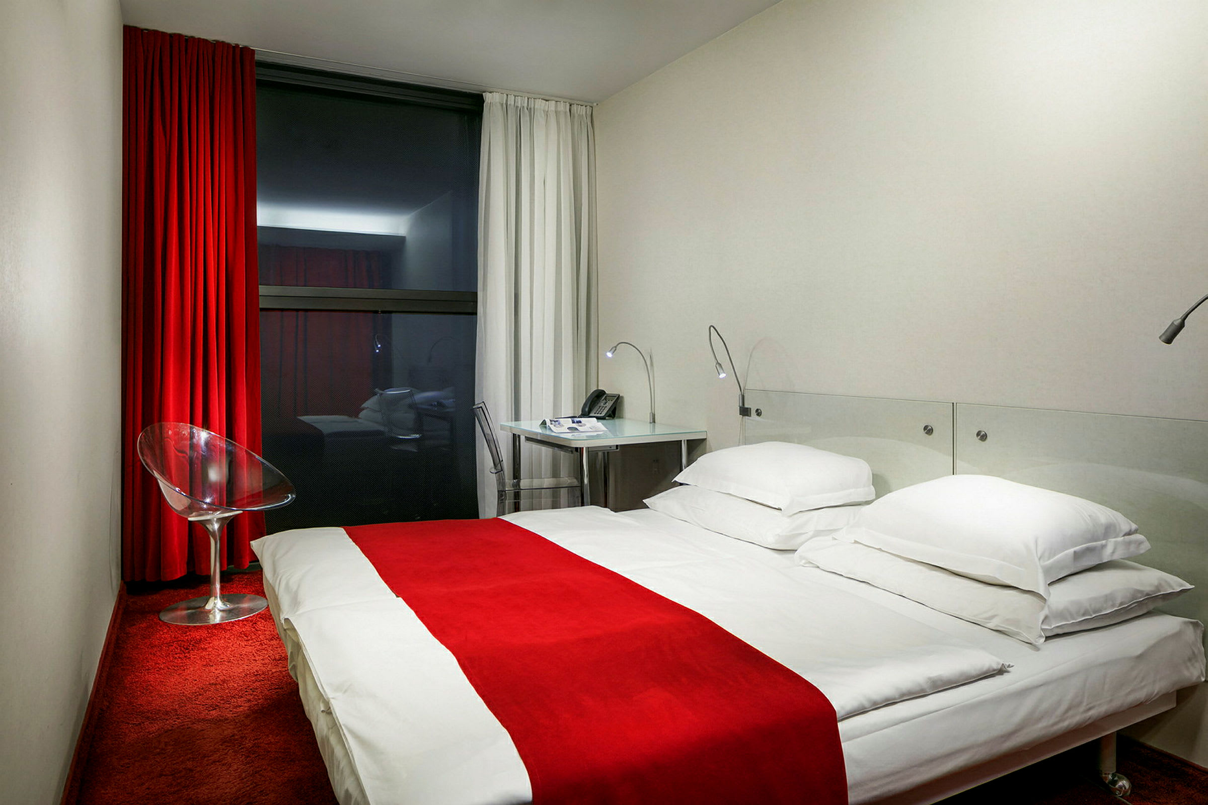 Hotelværelse med rødt tæppe, hvid seng med rødt sengetæppe, glasstol og skrivebord. (AI-genereret tekst)