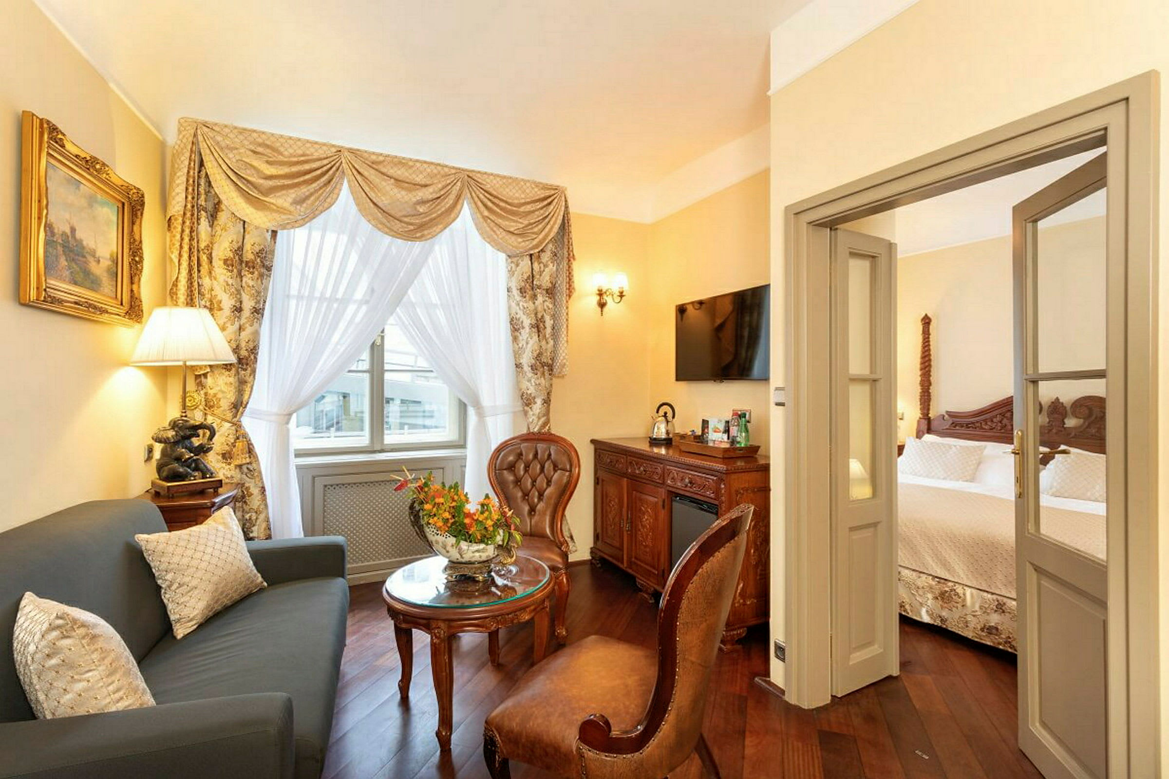 Et elegant indrettet hotelværelse med en stue med sofa og stole samt et soveværelse i baggrunden. Der er dekorative gardiner ved vinduet og en døråbning mellem rummene. (AI-genereret tekst)