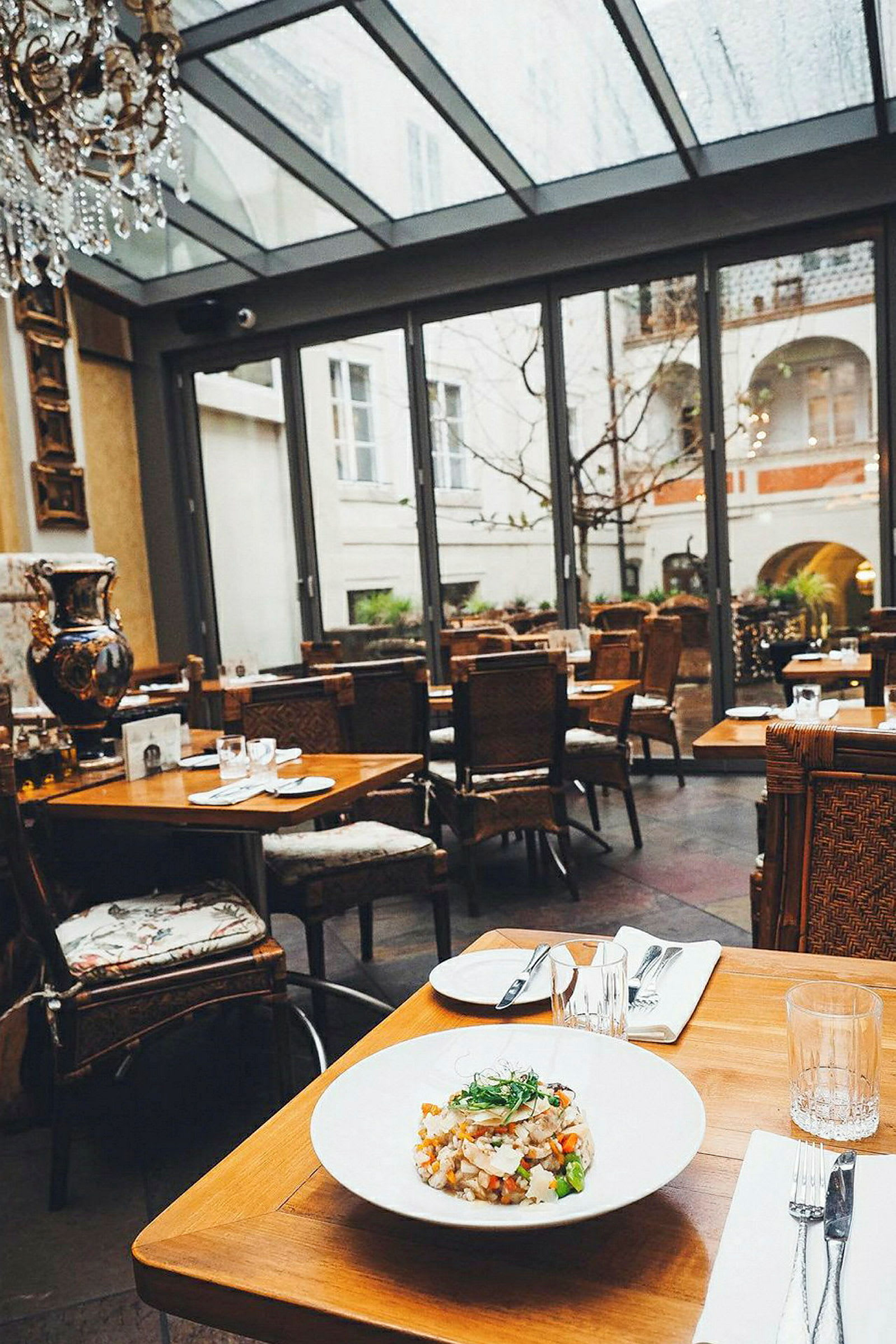 Et restaurantbord er dækket med en tallerken mad og bestik, placeret i et stilfuldt indrettet rum med store glasvinduer, der giver udsigt til en gårdhave. (AI-genereret tekst)