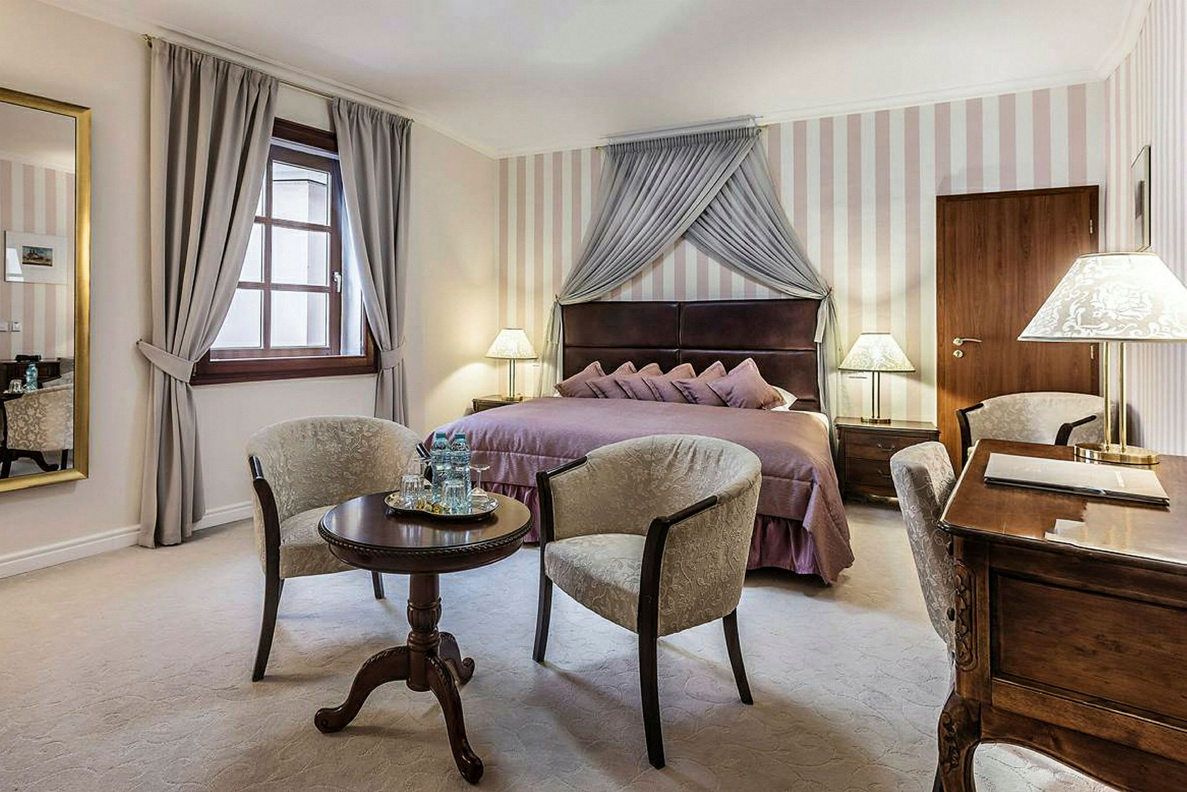 Et elegant hotelværelse med en dobbeltseng dækket med lilla sengetøj, to lænestole placeret rundt om et lille rundt bord og et skrivebord med lampe. (AI-genereret tekst)
