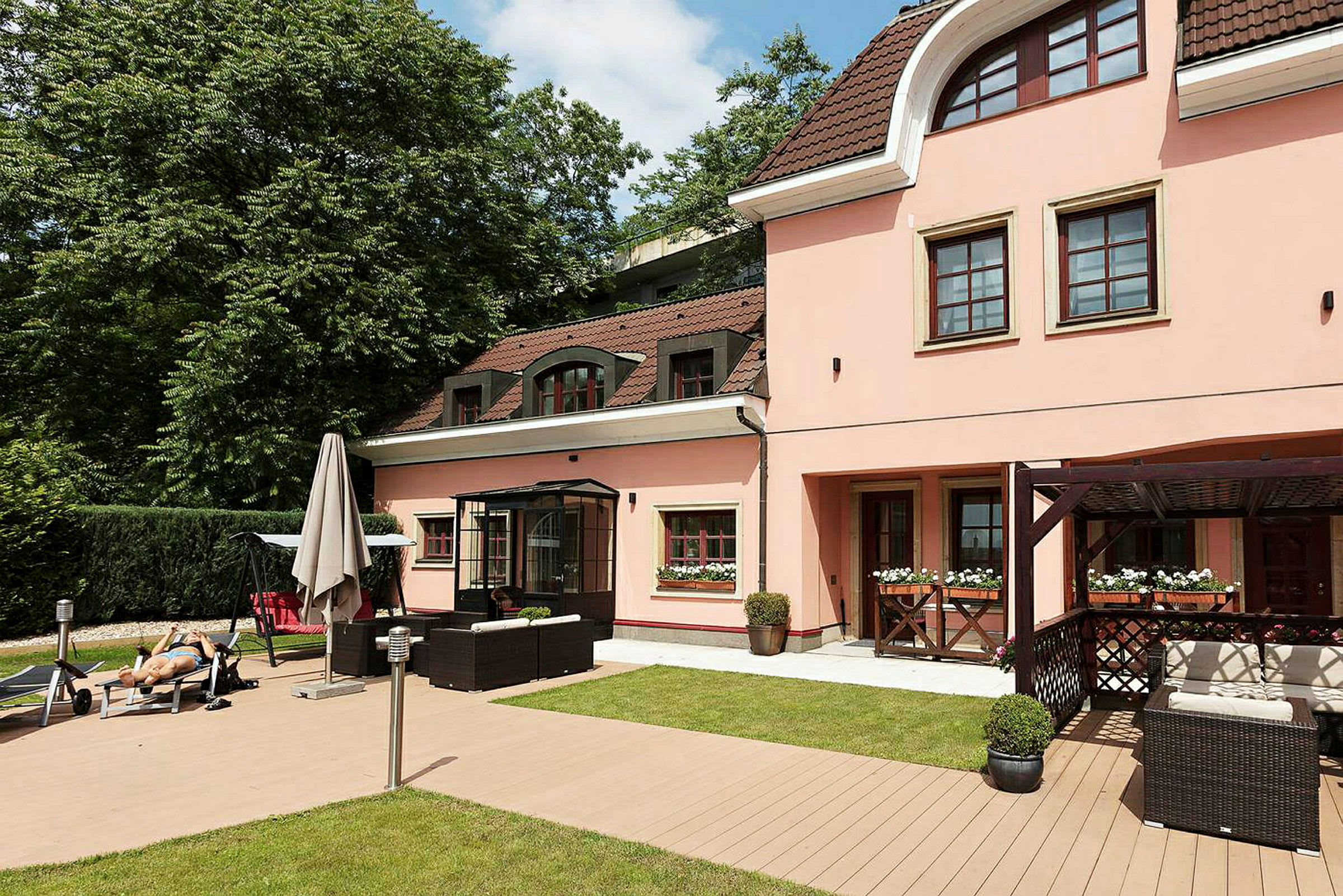 Et lyserødt hus med brunt tag og hvide vinduer, omgivet af en grøn have med møblerede terrasser og en terrasse. (AI-genereret tekst)
