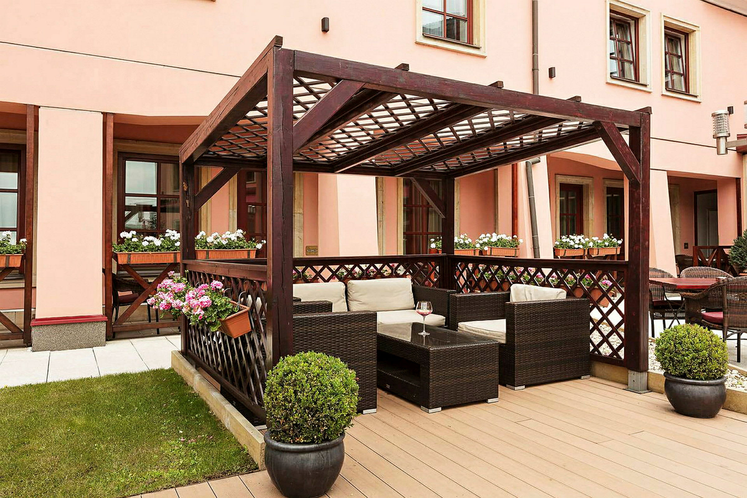 Billedet viser en terrasse med en pergola i mørkt træ, møbleret med kurvestole og borde. Blomsterkrukker med farverige blomster pryder området. (AI-genereret tekst)