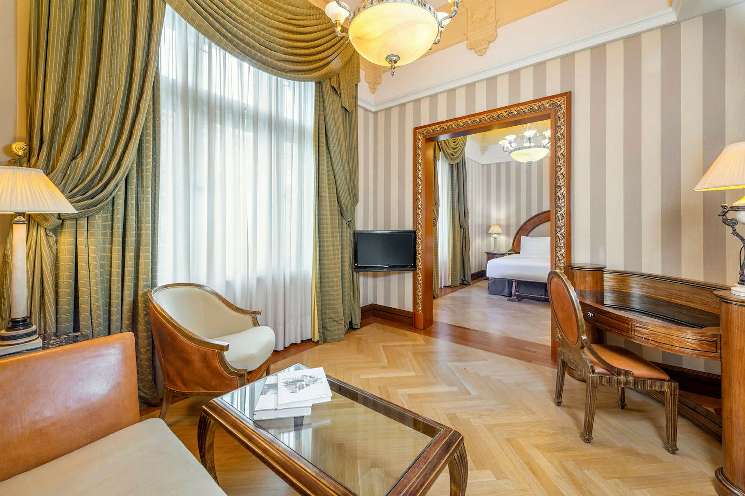 Et elegant hotelværelse med en siddegruppe bestående af stole og bord i forgrunden, samt en åben dør til soveværelset med en bred seng. (AI-genereret tekst)