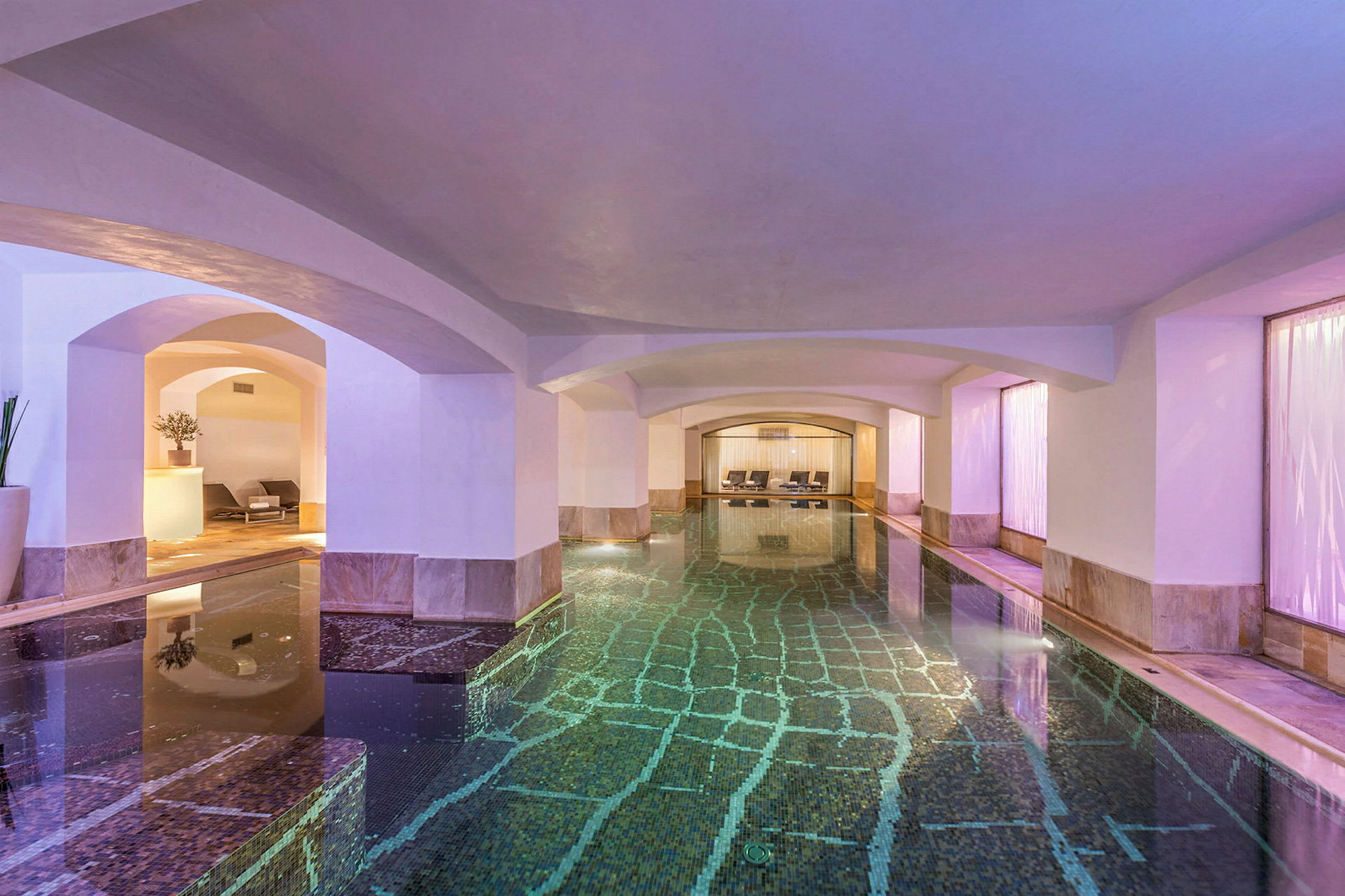 En indendørs pool med krystalklart vand omgivet af hvælvinger og moderne lounge møbler. (AI-genereret tekst)