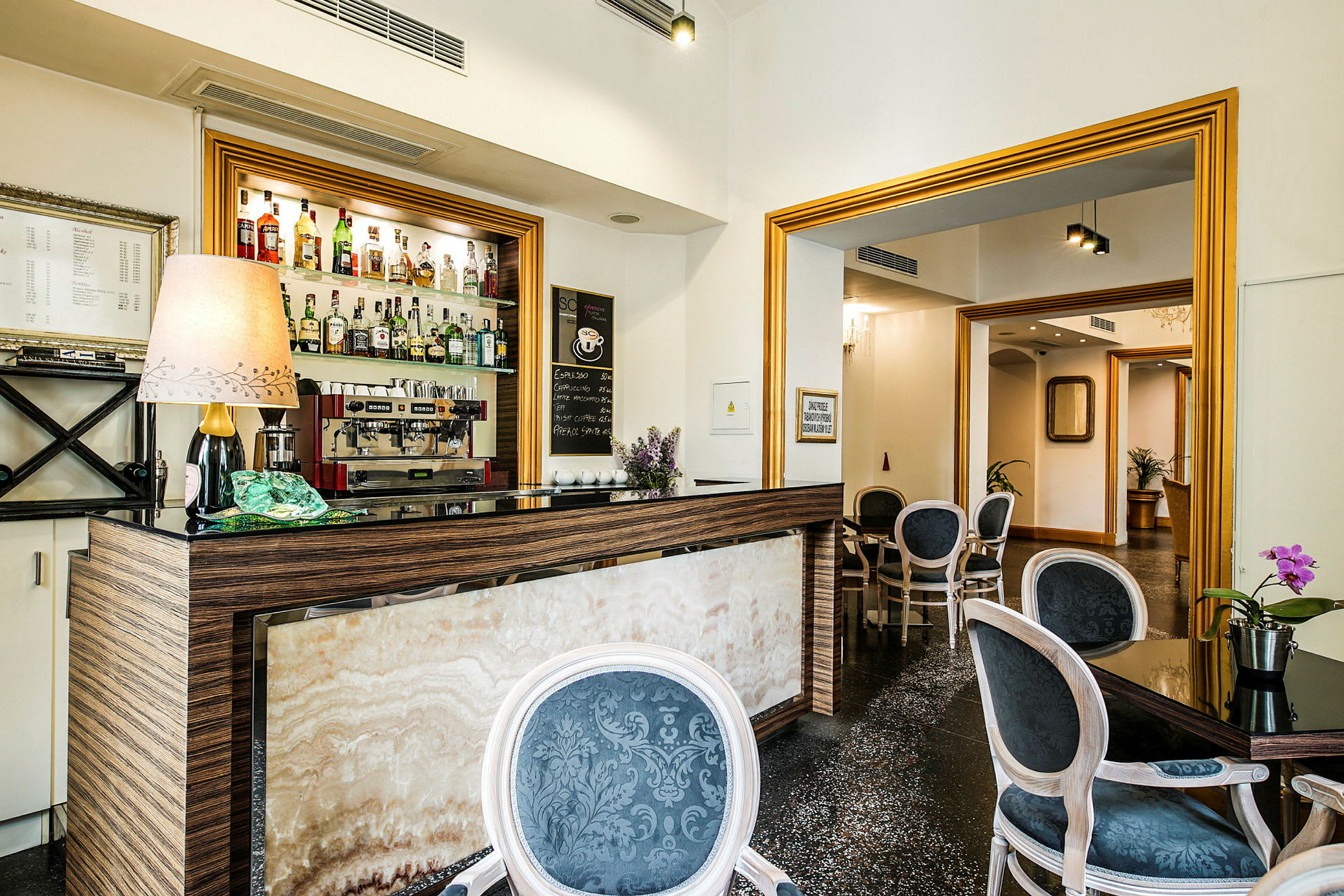 Billede af en lille hotelbar med forskellige typer spiritus på hylden samt en kaffemaskine, omgivet af møblerede siddepladser. (AI-genereret tekst)