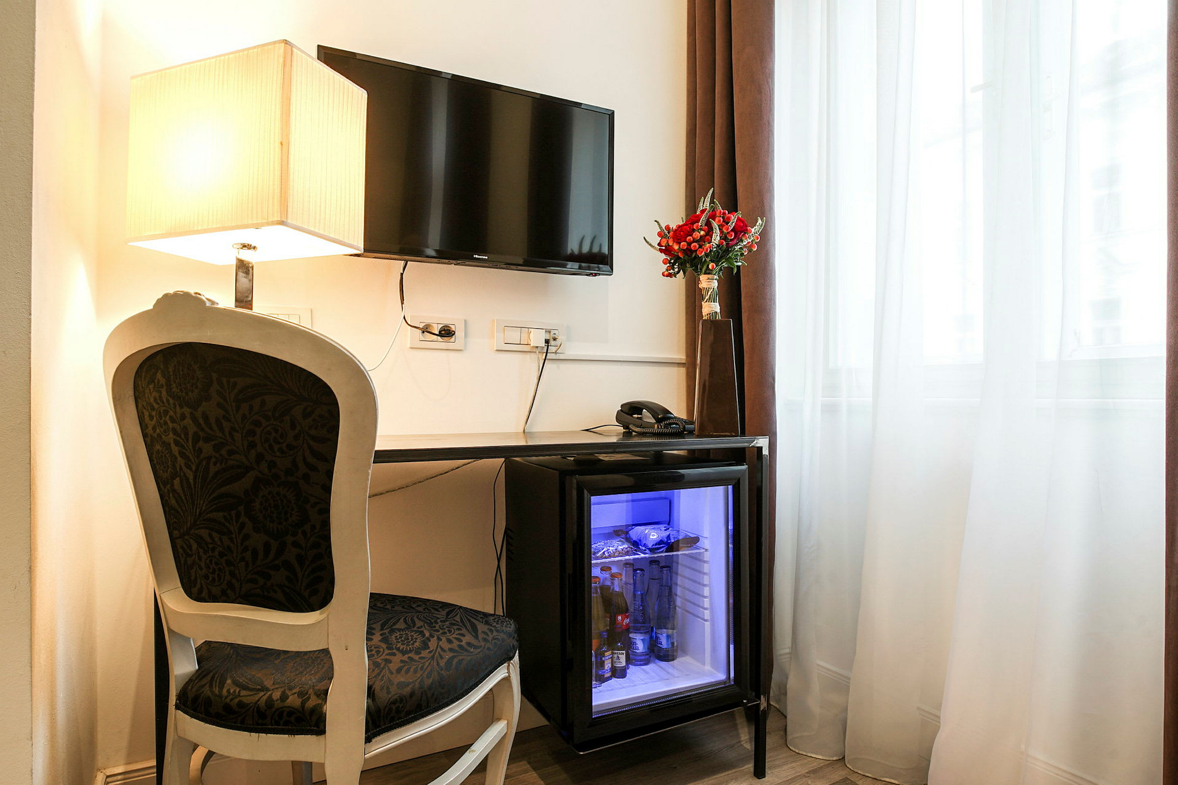 Et skrivebordsområde med en stol og et bord. Bordet har en minibar med glasdør, en telefon og en vase med røde blomster. Et vindue med gardiner og en vægmonteret TV samt en bordlampe. (AI-genereret tekst)