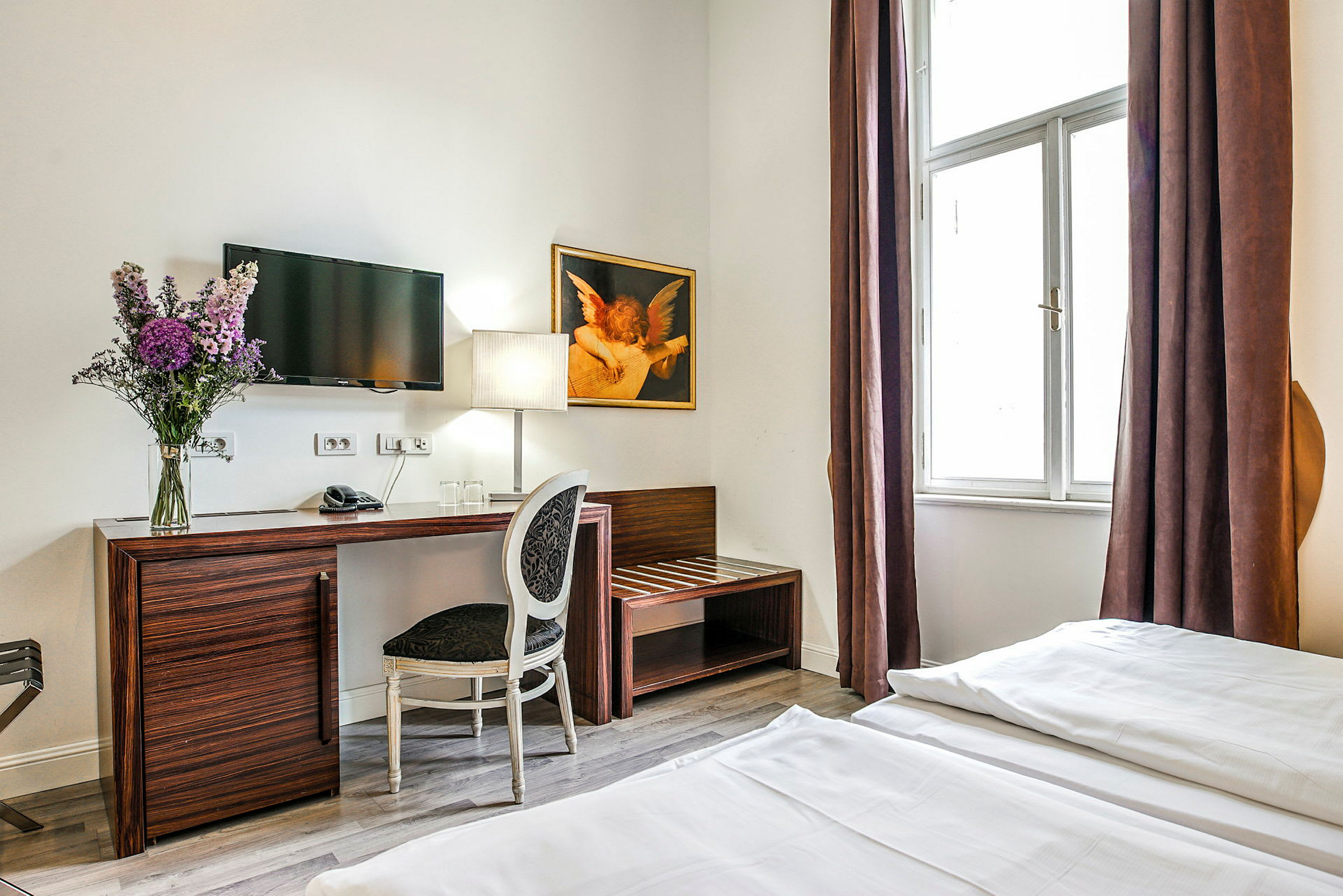 Et hotelværelse med et skrivebord og en stol, en tv på væggen, en blomsterbuket og en lampe placeret på bordet samt et stort vindue med brune gardiner. (AI-genereret tekst)