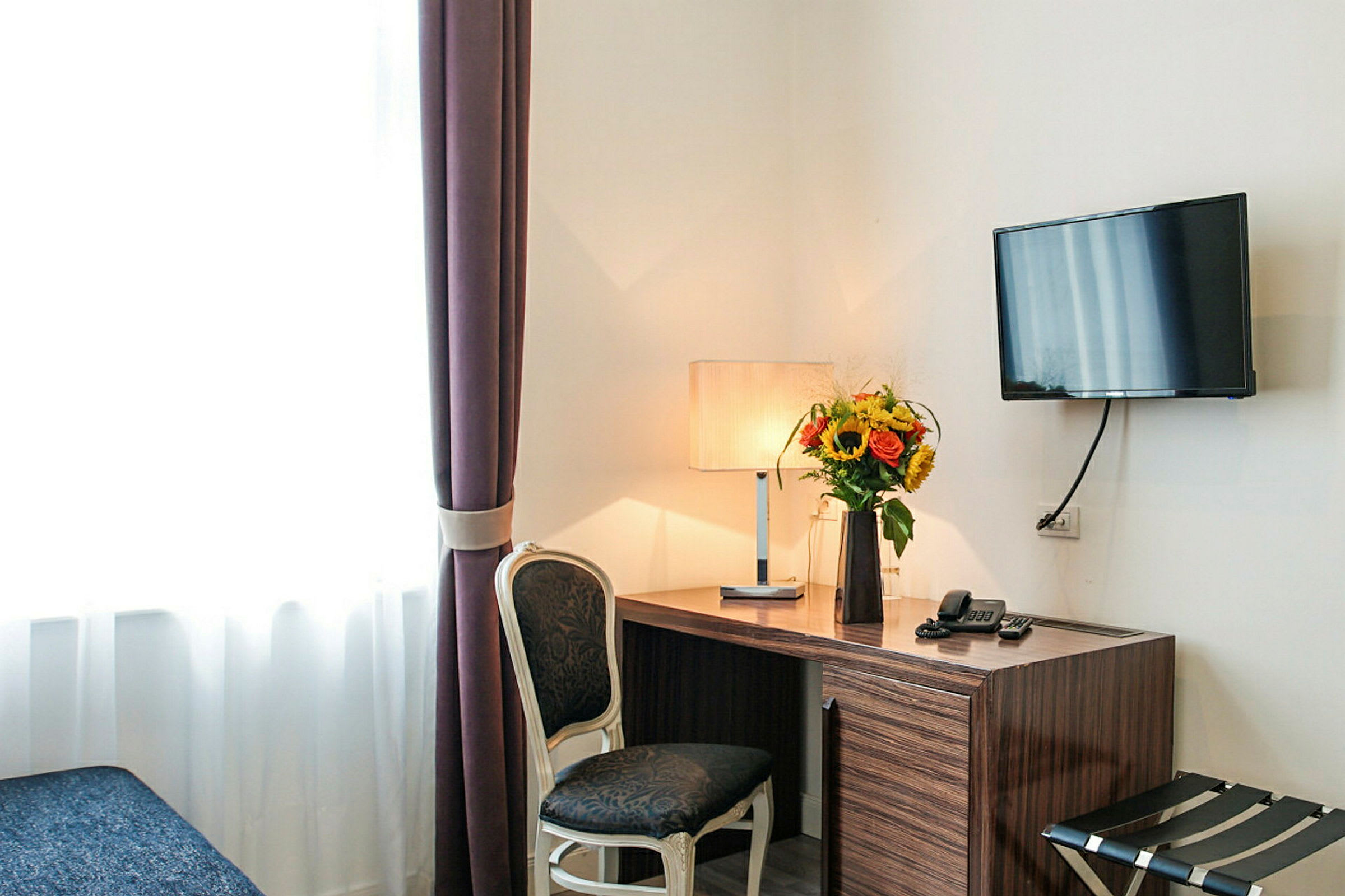 Et skrivebord med en lampe og en vase med blomster placeret ovenpå, ved siden af en stol og et vægmonteret fjernsyn. (AI-genereret tekst)