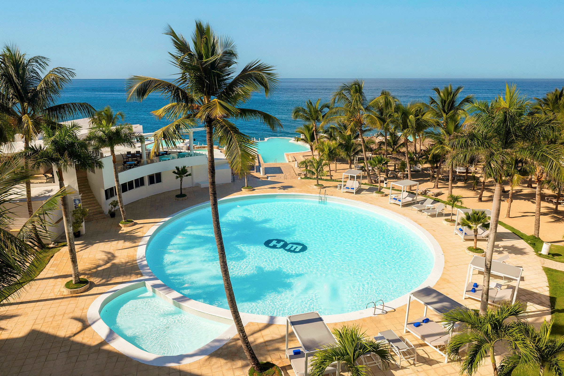 Et hotelområde med en stor rund pool omgivet af palmer, solsenge og cabanaer. Havudsigt i baggrunden. (AI-genereret tekst)