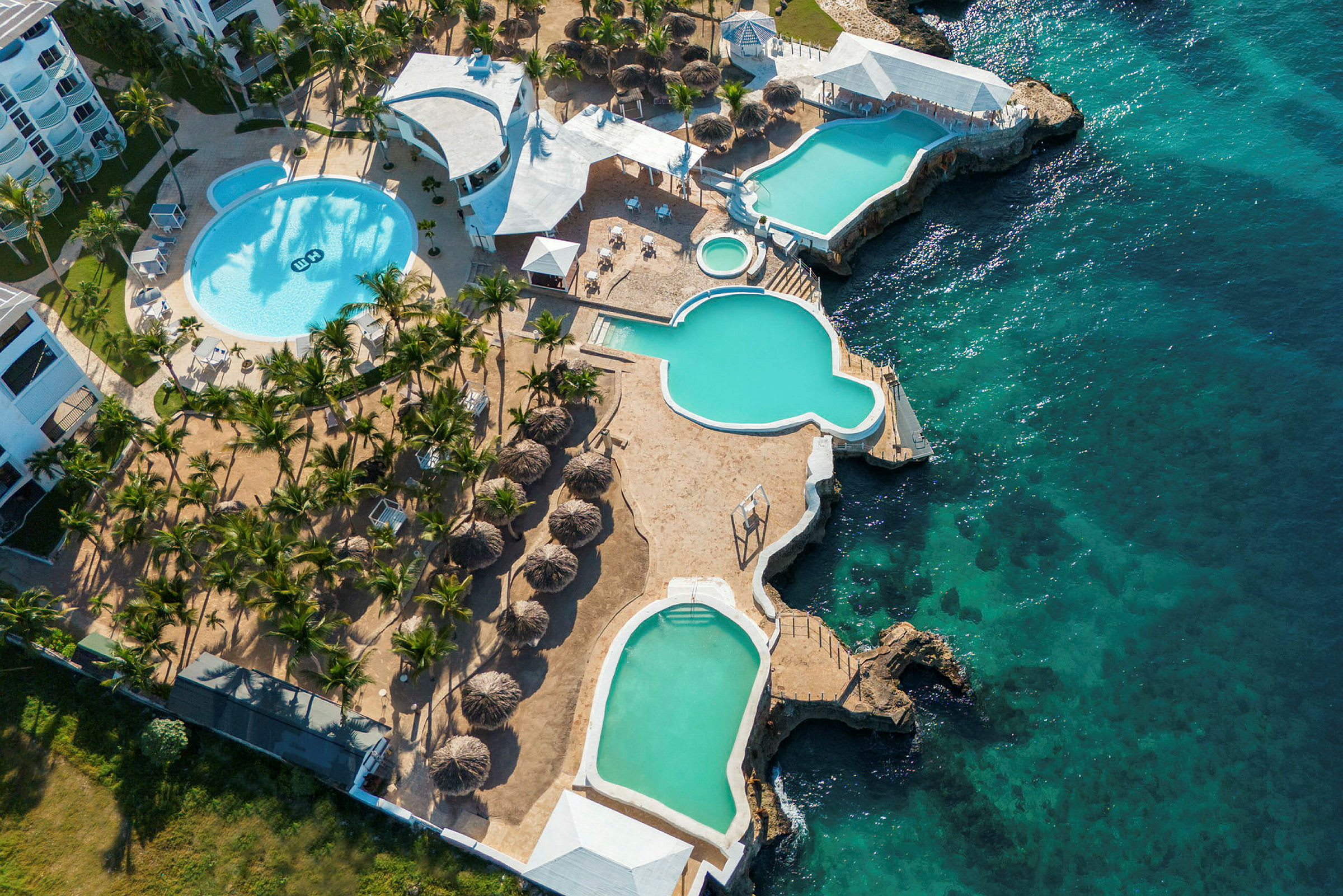 Luftfoto af et resort med flere pools, omgivet af solsenge og palmer, ved siden af et klart blåt hav. (AI-genereret tekst)