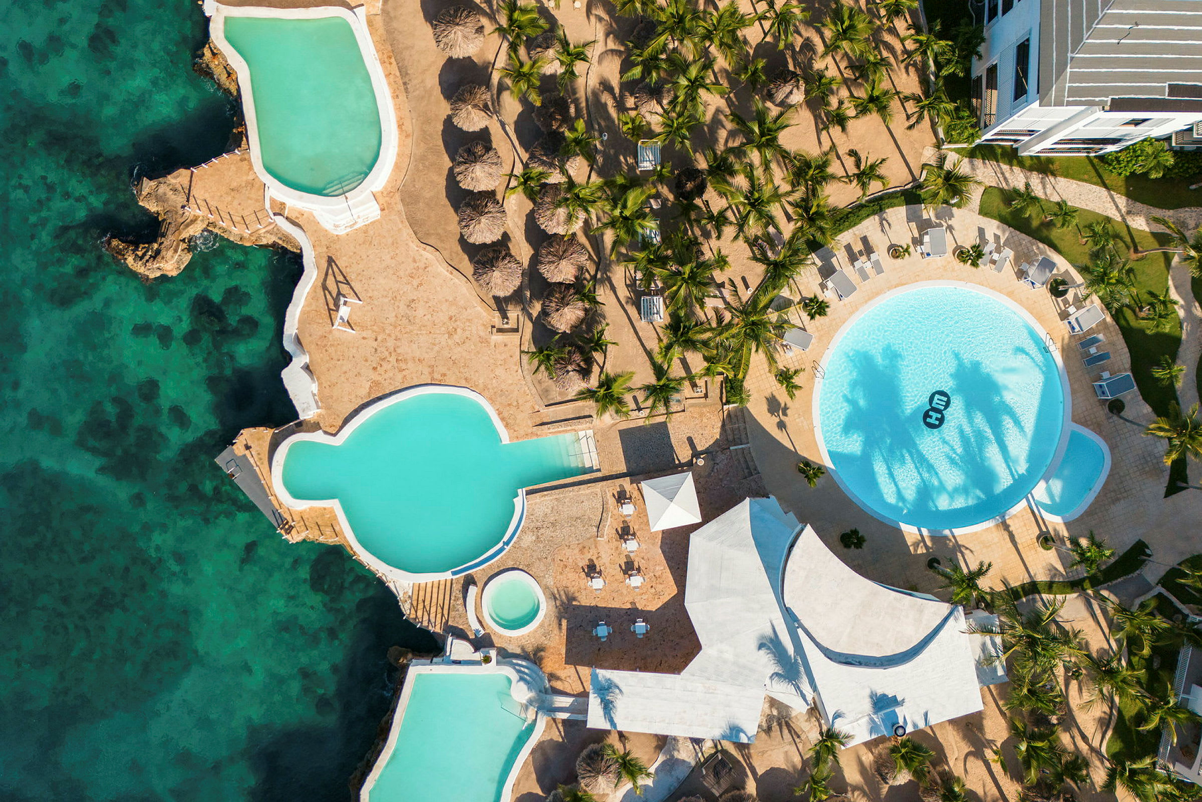 Luftfoto af et feriested med swimmingpools, palmer og hav. (AI-genereret tekst)