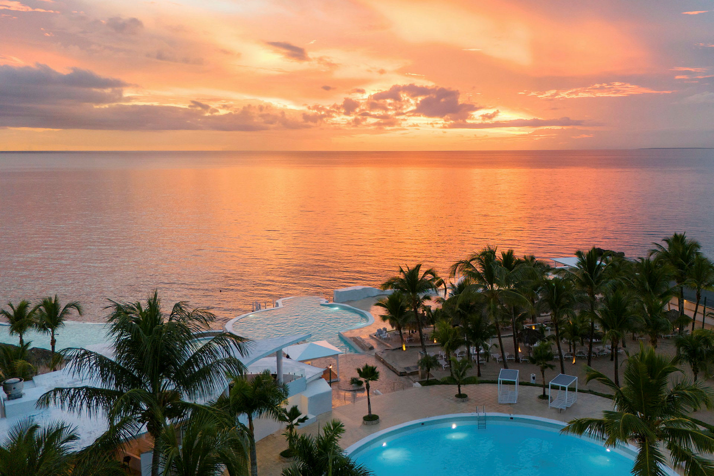 En udsigt over et feriested med swimmingpools og palmer ved havet under en farverig solnedgang. (AI-genereret tekst)