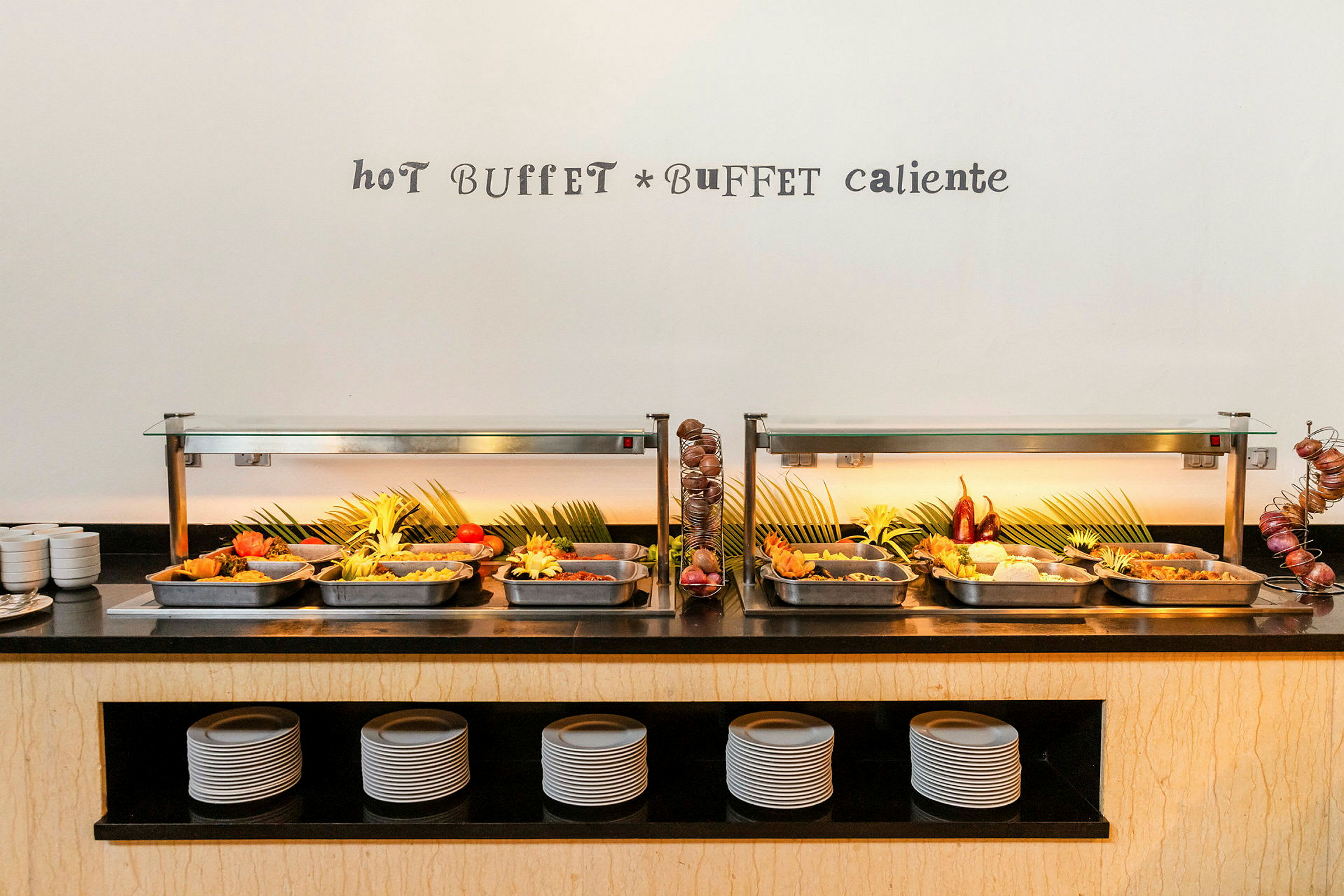 En varm buffet med forskellige retter arrangeret i stålforme og dekoreret med grønne blade og friske grøntsager. (AI-genereret tekst)