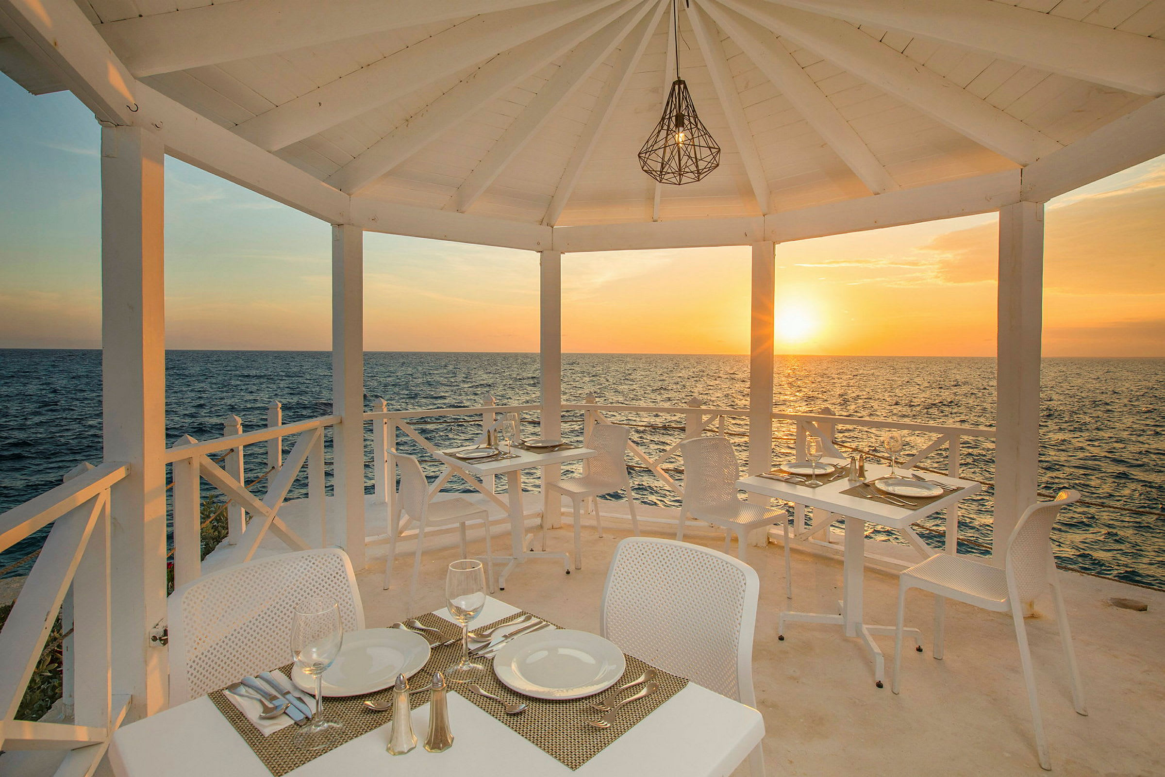 En udendørsrestaurant med hvide møbler, beliggende ved havet, med solnedgang i baggrunden. (AI-genereret tekst)
