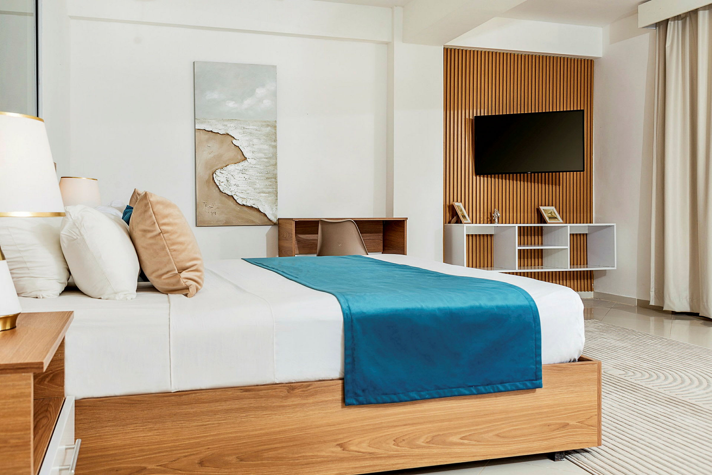 Et soveværelse med en stor træbed, hvide og beige puder og et blåt sengetæppe. Et billede med strandmotiv hænger på væggen, og et TV er monteret på en træpanel. Et skrivebord med stol er ved siden af. (AI-genereret tekst)