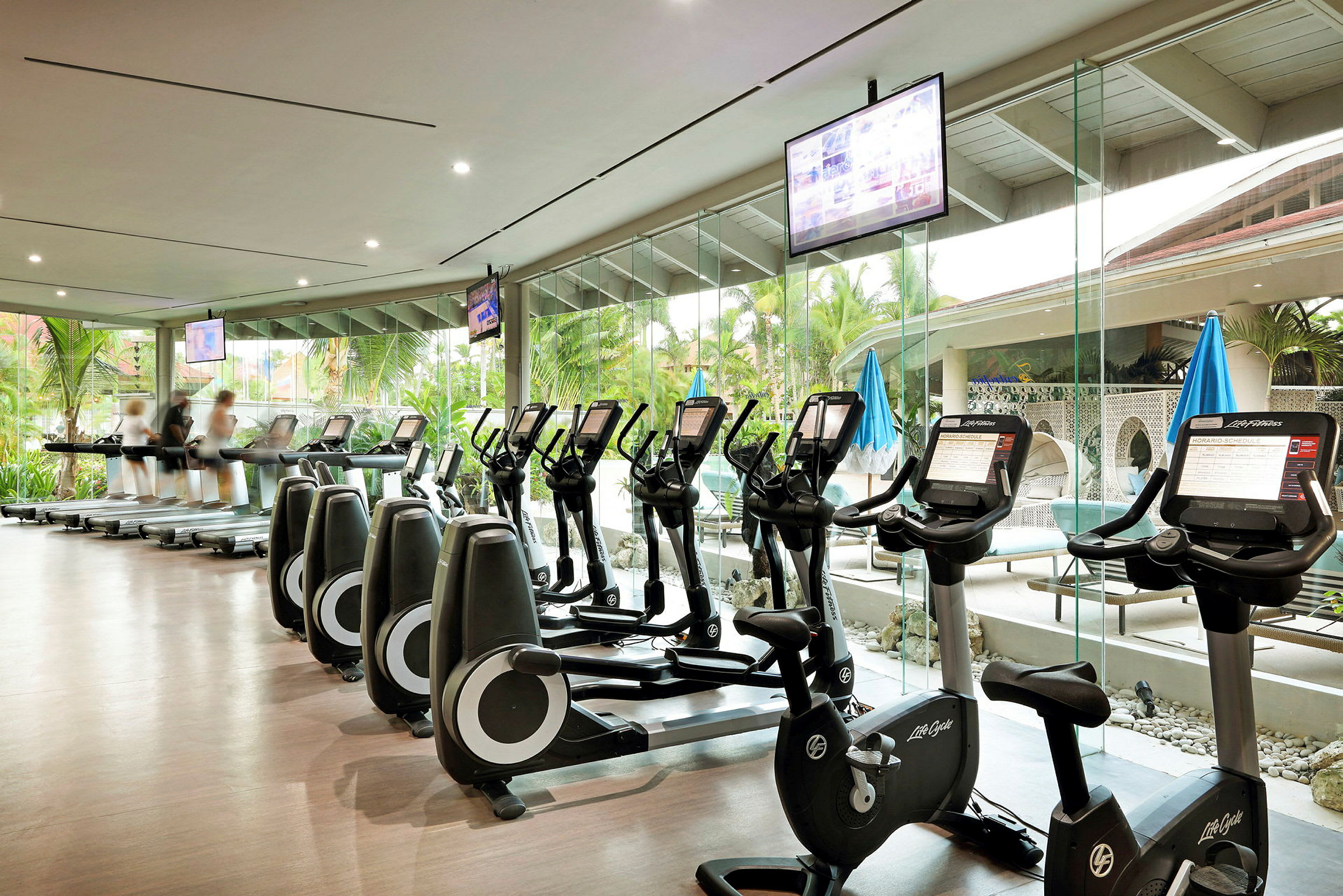 Et fitnesscenter med træningsmaskiner, herunder løbebånd og motionscykler. Store vinduer viser grønne områder og en udendørs miljø. (AI-genereret tekst)