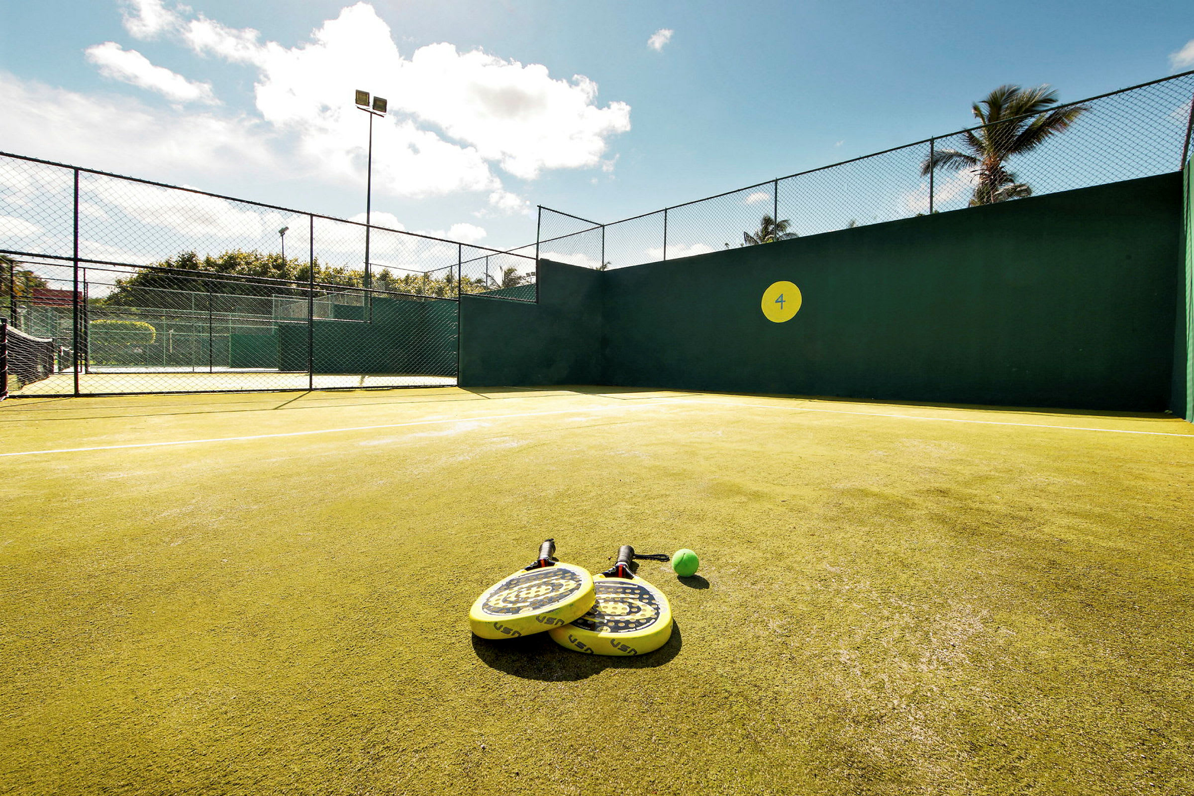 En padelbane med gule ketsjere og en bold placeret på jorden. Solrigt vejr. (AI-genereret tekst)