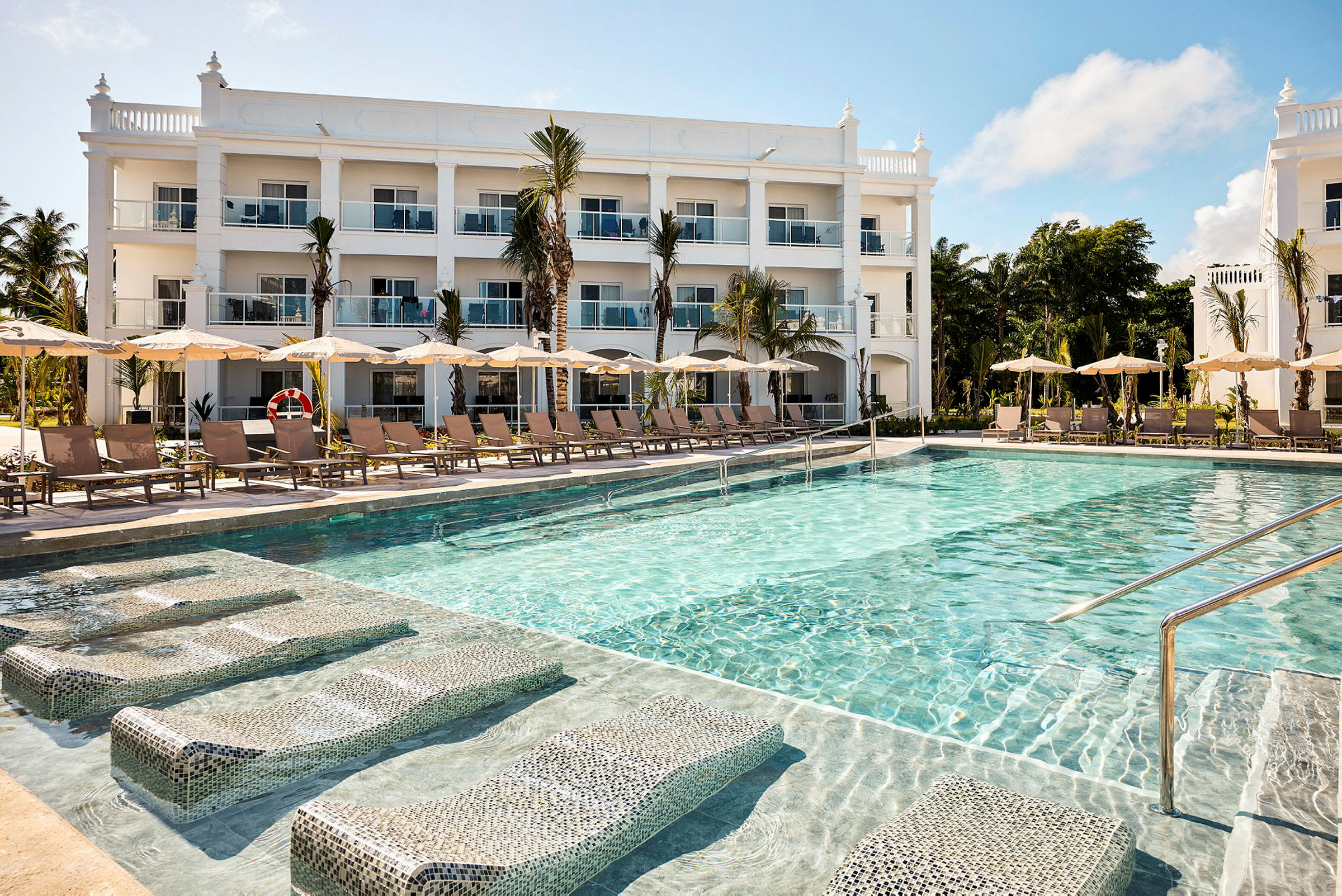 En swimmingpool foran et hotel med hvid facade og flere etager. (AI-genereret tekst)