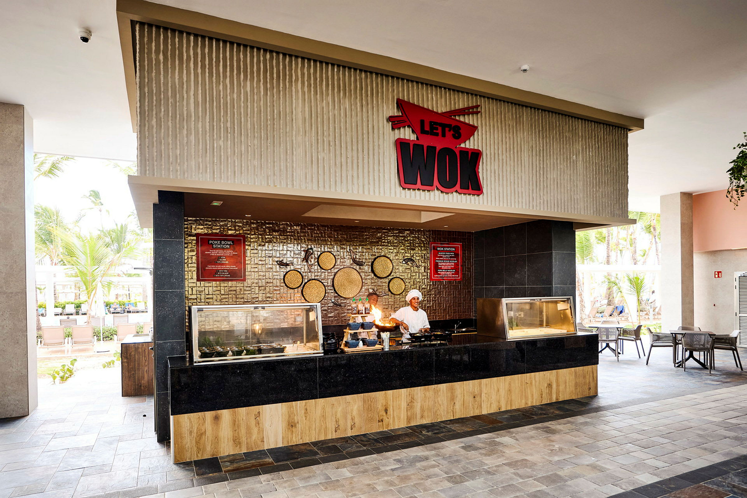 Billede af en restaurant med en serveringsdisk markeret 'Let's Wok'. Interiøret har et moderne design med træ og sorte paneler, og der er udendørs omgivelser med palmer i baggrunden. (AI-genereret tekst)