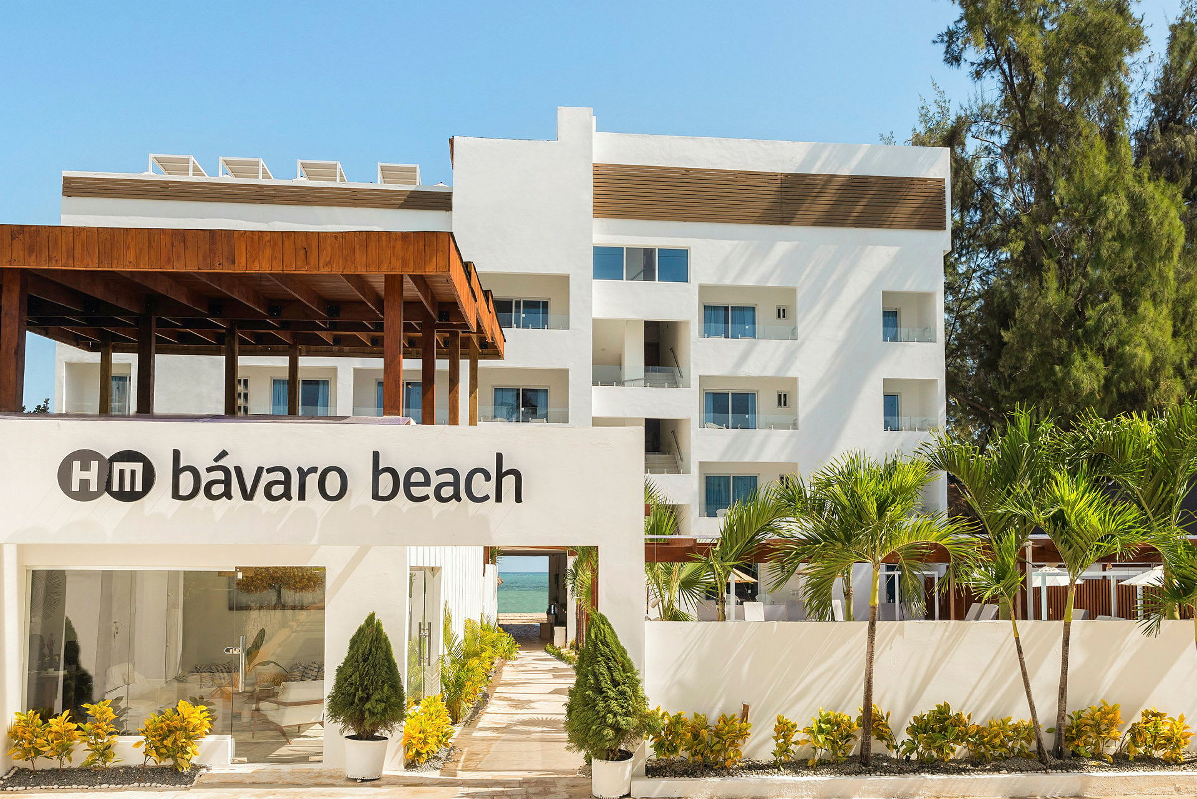 Billede af en hvid bygning med teksten "HM Bávaro Beach" på en væg foran, omgivet af frodig grøn bevoksning og palmer. (AI-genereret tekst)