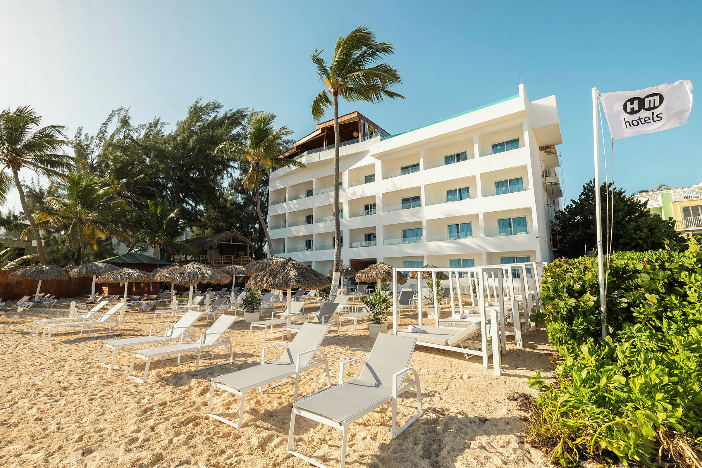 En hvid hotelbygning ved en sandstrand omgivet af parasoller og liggestole. (AI-genereret tekst)