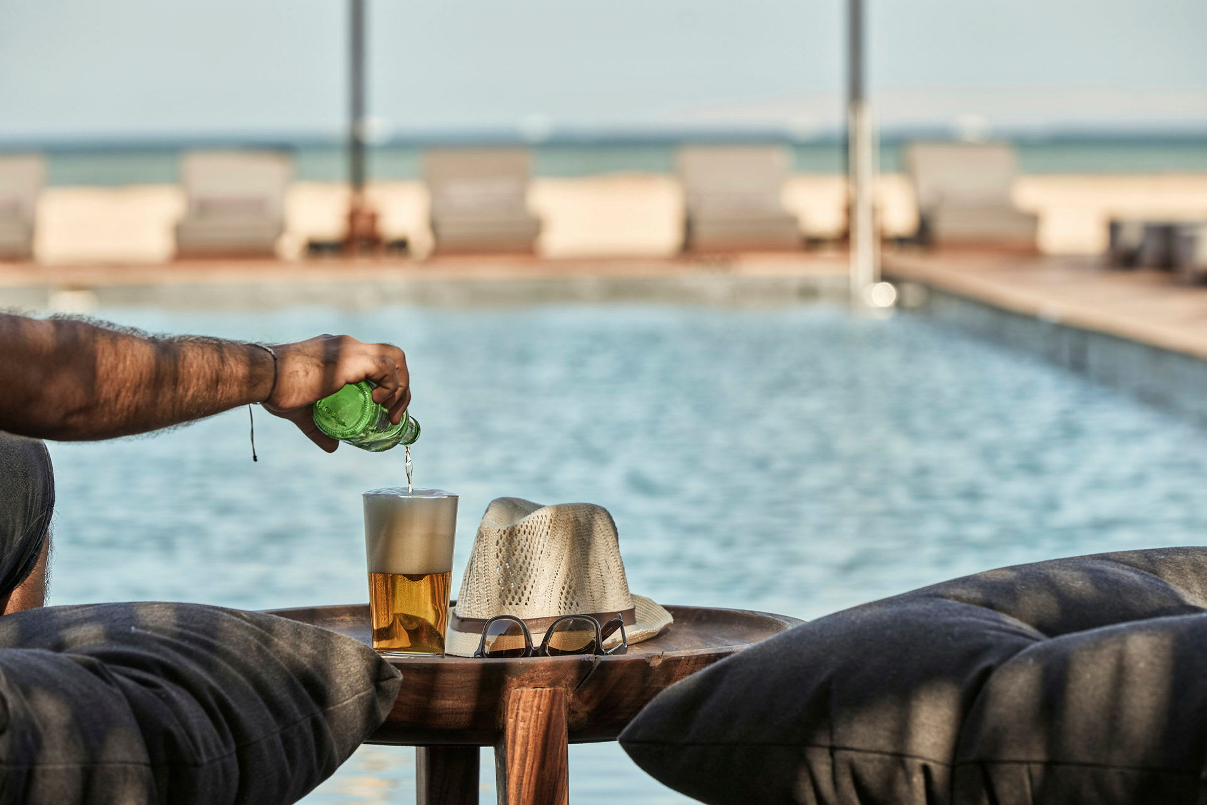 En person hælder drikke fra en grøn flaske i et glas ved poolen, med en hat og briller på et bord. (AI-genereret tekst)