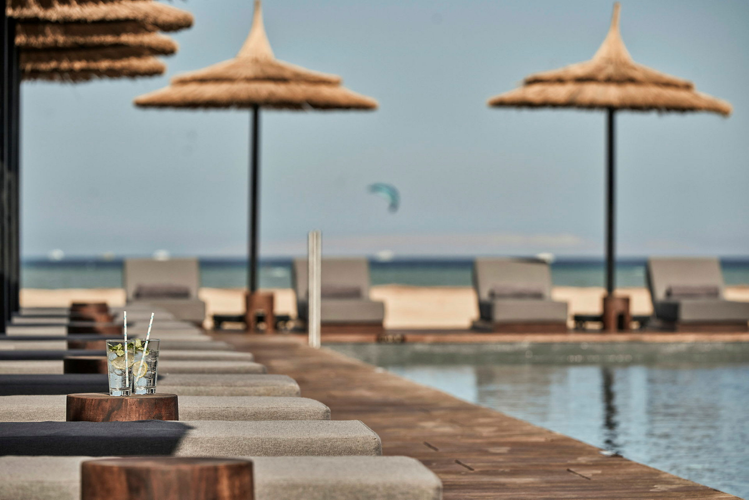 Liggestole med parasoller ved en pool, med stranden og havet i baggrunden. (AI-genereret tekst)