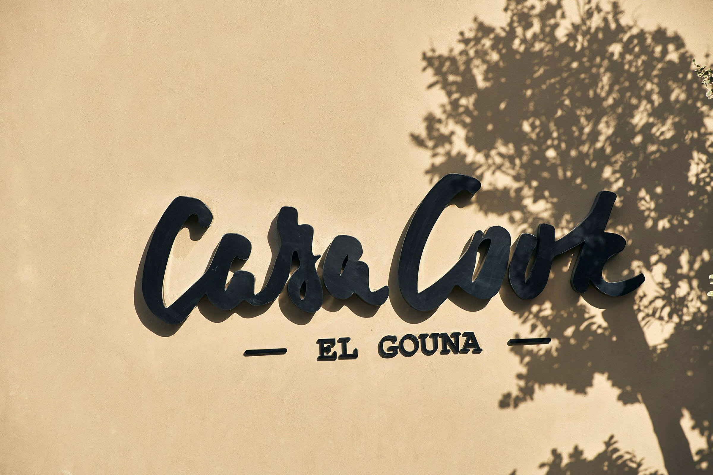 Logoen for Casa Cook El Gouna på en væg, med skyggen af et træ. (AI-genereret tekst)