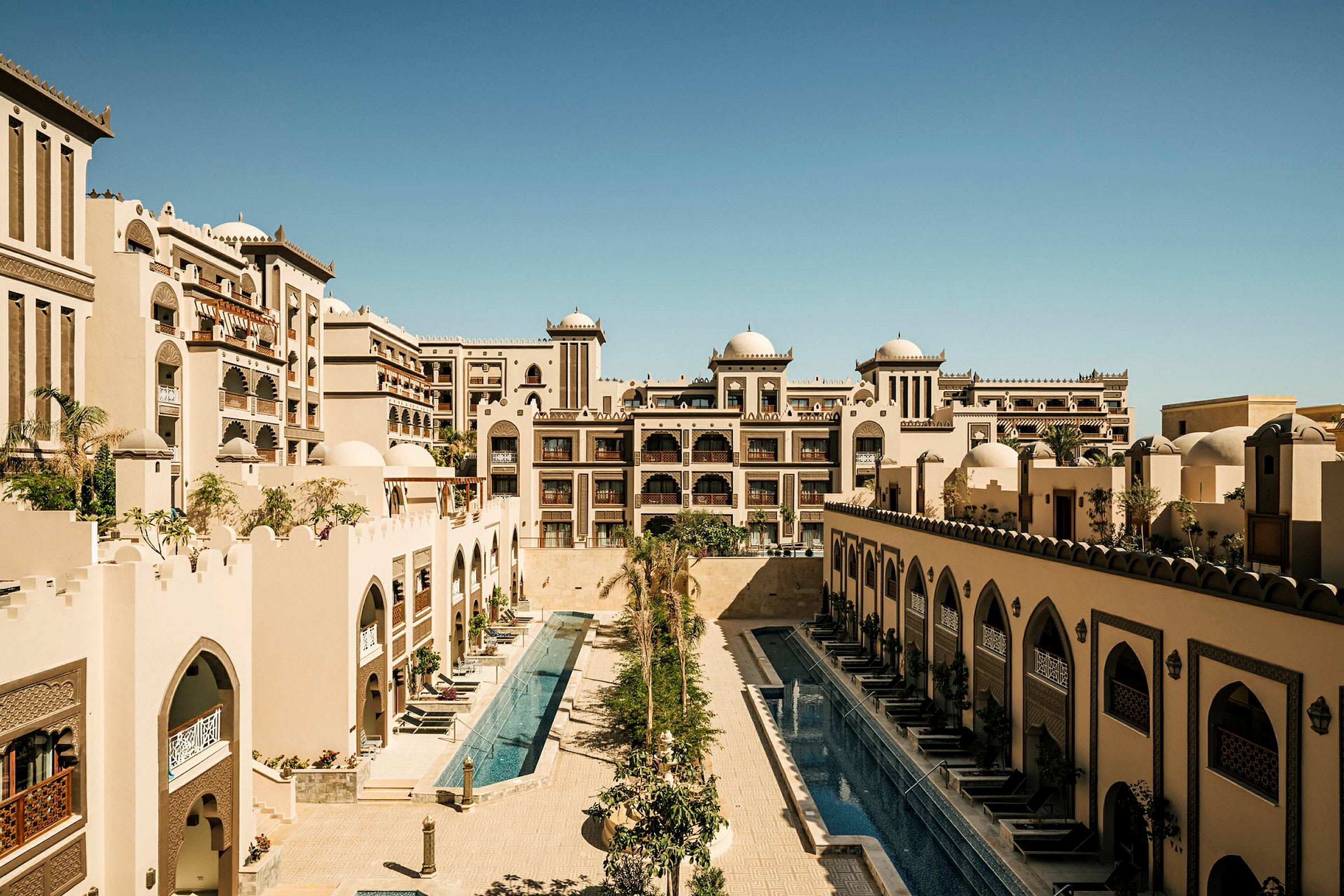 Billedet viser et hotelkompleks med arkitektur inspireret af arabisk stil, herunder kupler og ornamenterede buer, samt to aflange swimmingpools omgivet af bygninger. (AI-genereret tekst)