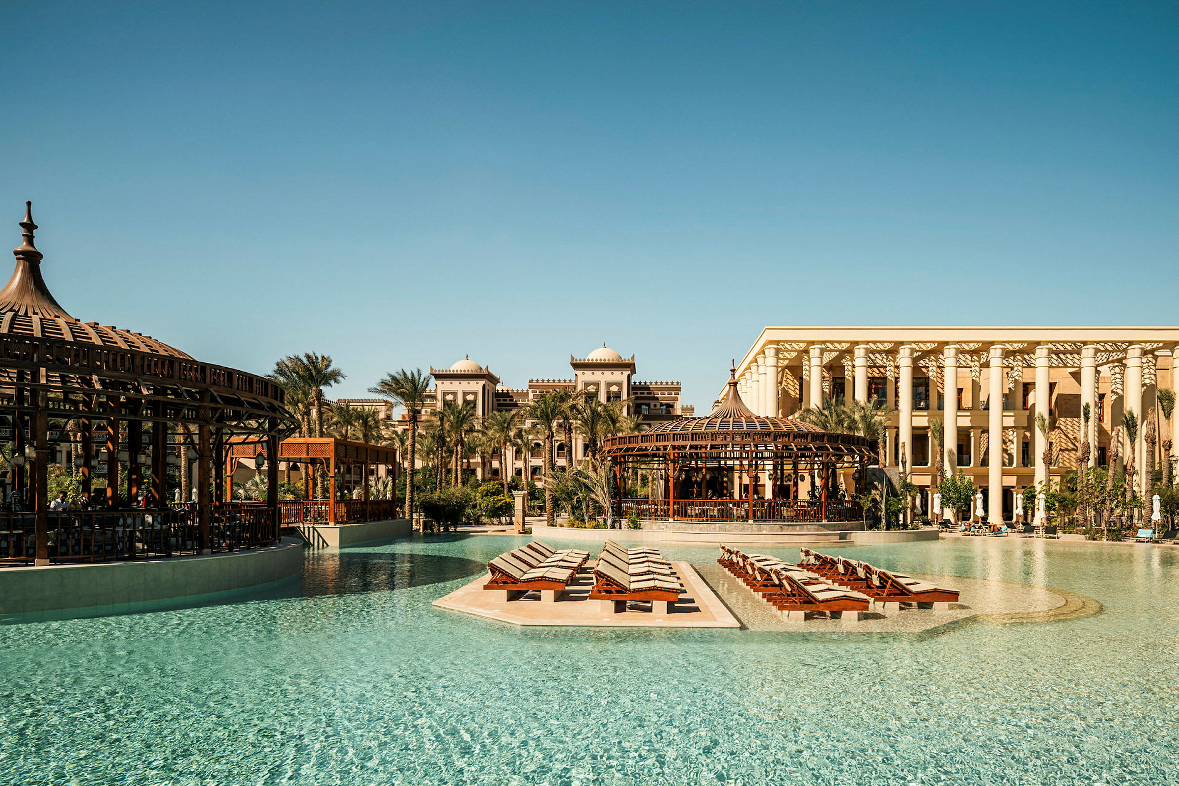 Et postkort-lignende udendørsbillede af et luksushotel med pool og liggestole omgivet af palmer og traditionel arkitektur. (AI-genereret tekst)