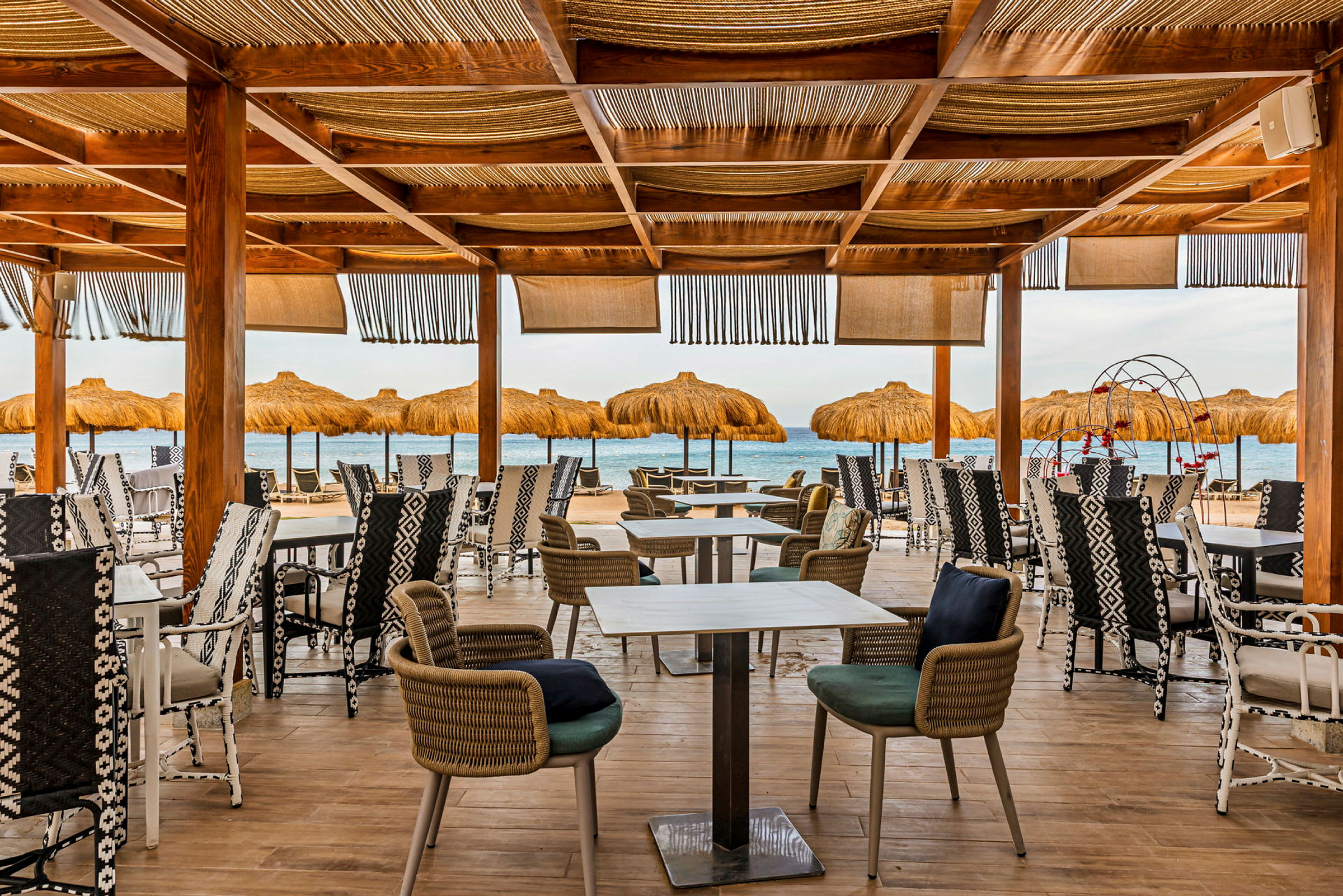 En udendørs restaurant med træstolper og stole, med udsigt over havet og parasoller på stranden. (AI-genereret tekst)