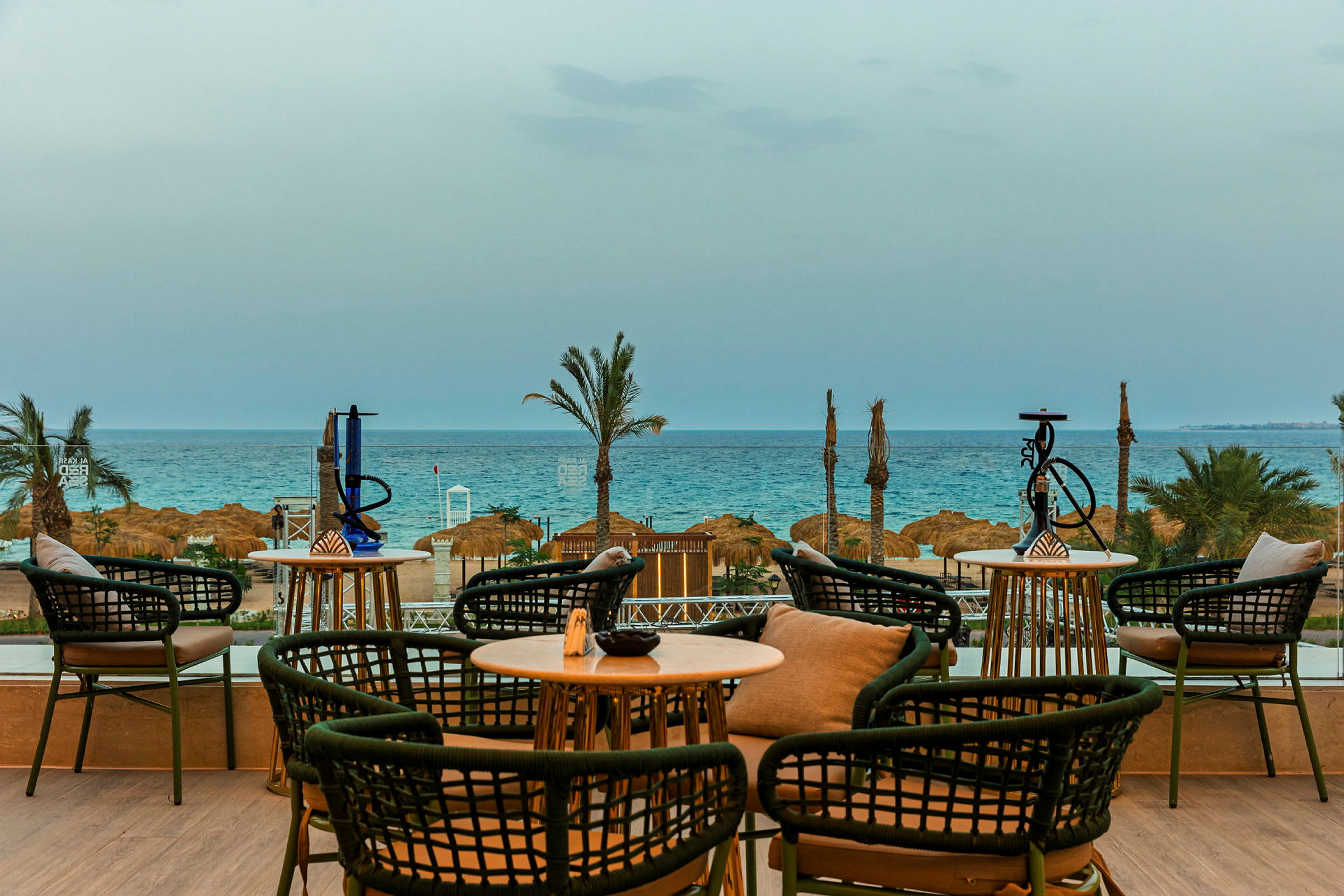 En udsigt fra en terrasse med siddepladser, borde og vandpiber, der vender ud mod stranden og havet i baggrunden. (AI-genereret tekst)