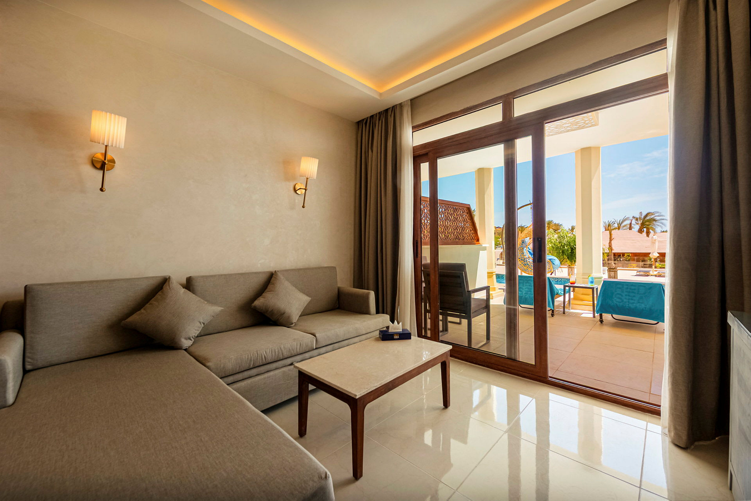 En hotelsuite med en grå sofa, et sofabord og to væglamper. Udenfor glasdørene ses en terrasse med siddepladser og solsenge ved en pool. (AI-genereret tekst)