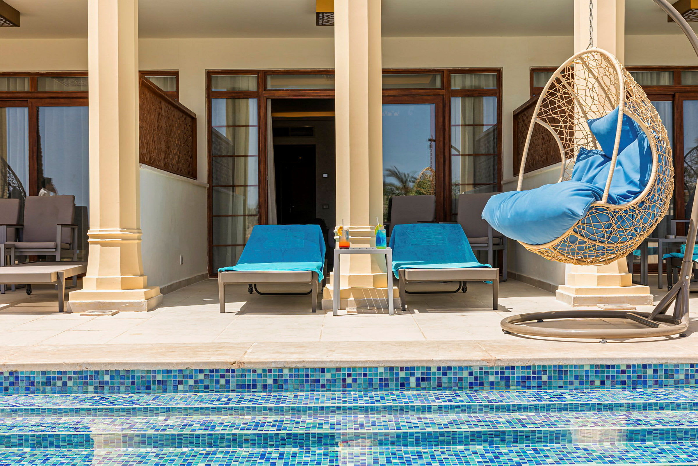 En terrasse med to liggestole med blå håndklæder, en rund rottinghængestol med blå pude og et bord med farverige drikkevarer. Foran terrassen er en swimmingpool. (AI-genereret tekst)