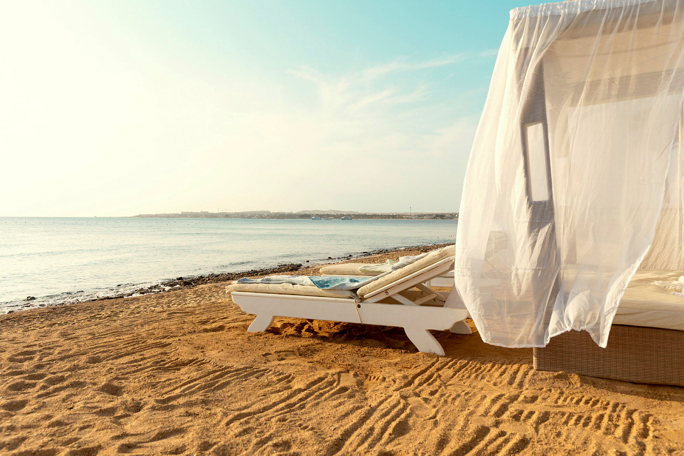 En solseng med et hvidt draperi på en sandstrand ved havet. (AI-genereret tekst)
