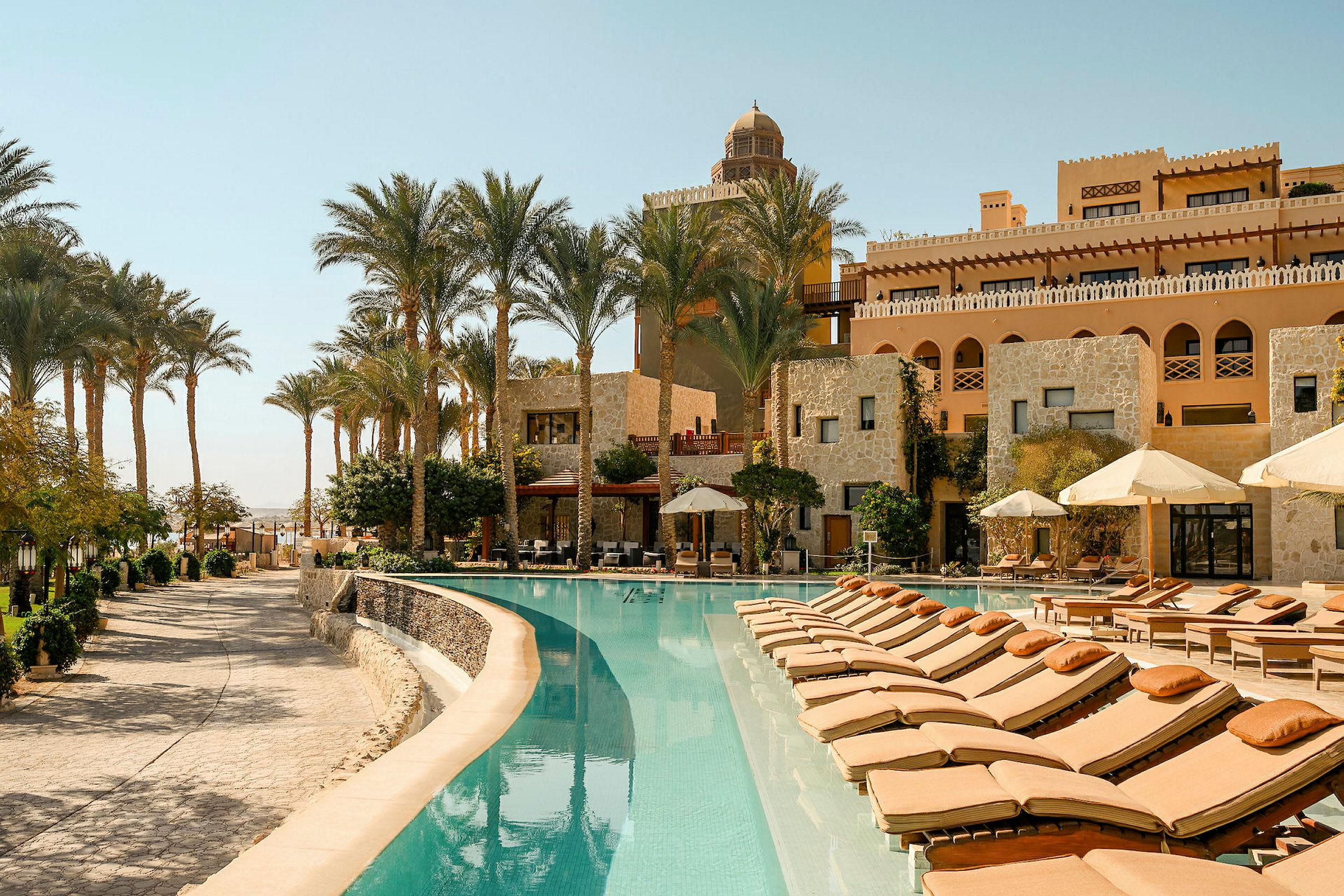 Et elegant hotelområde med en pool omgivet af liggestole og parasoller. Bygningerne i baggrunden er bygget i sten og dekoreret med buede vinduer. Palmer omgiver området. (AI-genereret tekst)