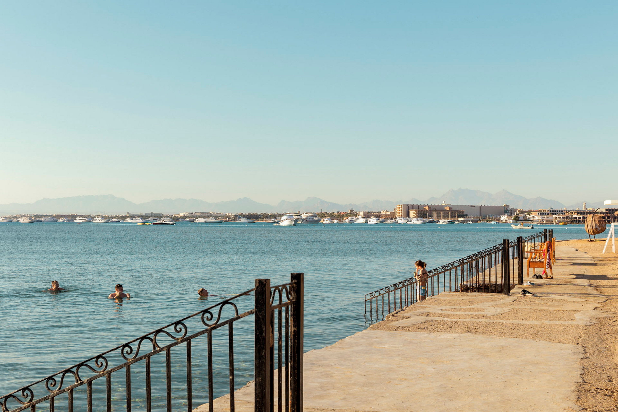En strandpromenade med personer, der svømmer i havet, både i det fjerne og bjerge i baggrunden. (AI-genereret tekst)