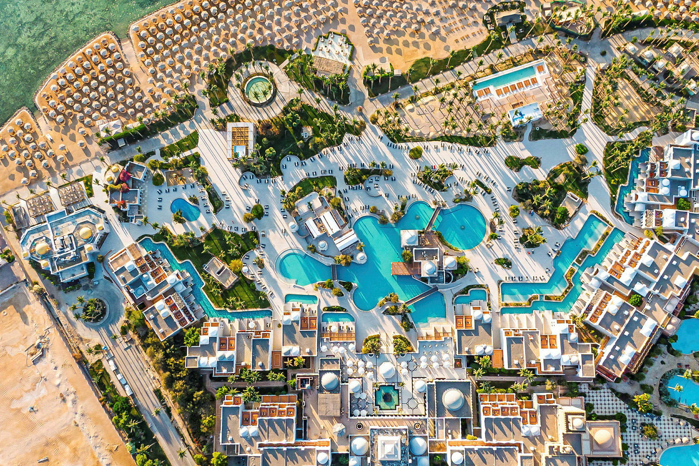 Luftfoto af et ferieområde med swimmingpools, bygninger og en strand med parasoller. (AI-genereret tekst)