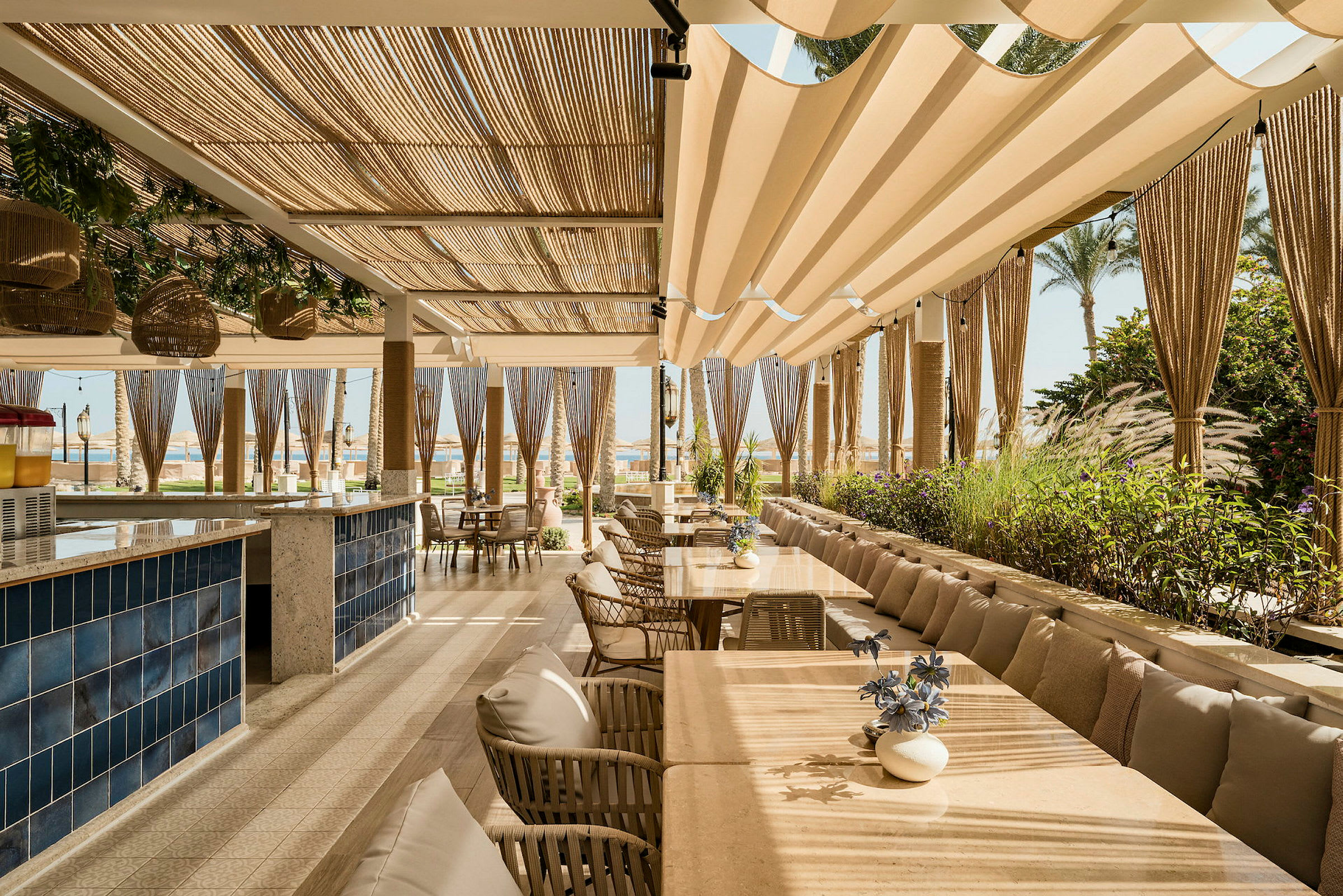 En restaurantterrasse med borde, stole og beige solsejl, omgivet af grønne planter. (AI-genereret tekst)