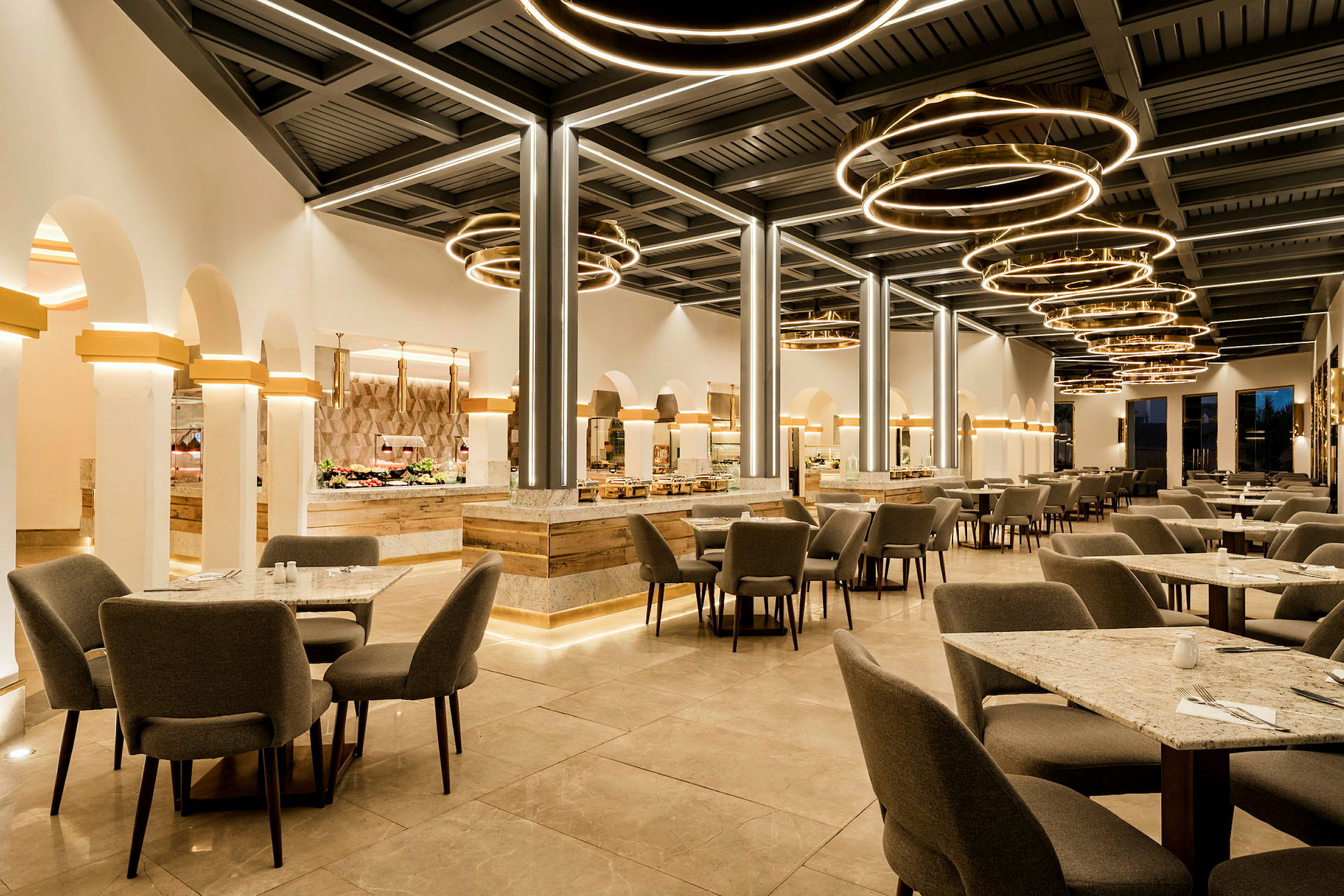 En moderne restaurant med grå stole og borde på et stenlagt gulv, lyse cirkelformede lamper hænger fra loftet. (AI-genereret tekst)