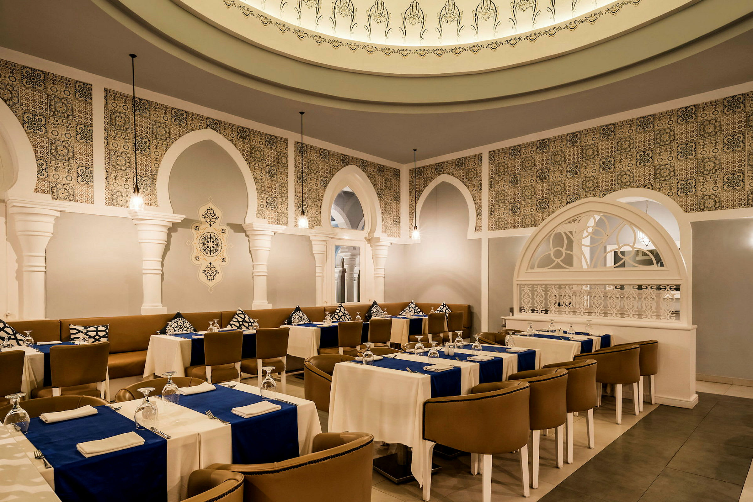 En elegant restaurant med hvide søjler, dekorativt flisemønster og dækkede borde med blå duge. (AI-genereret tekst)