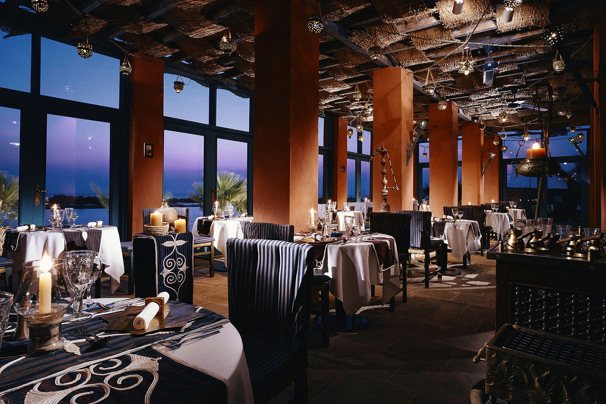 Restaurant med belysning, borde og stole samt store vinduer mod skumringen. (AI-genereret tekst)