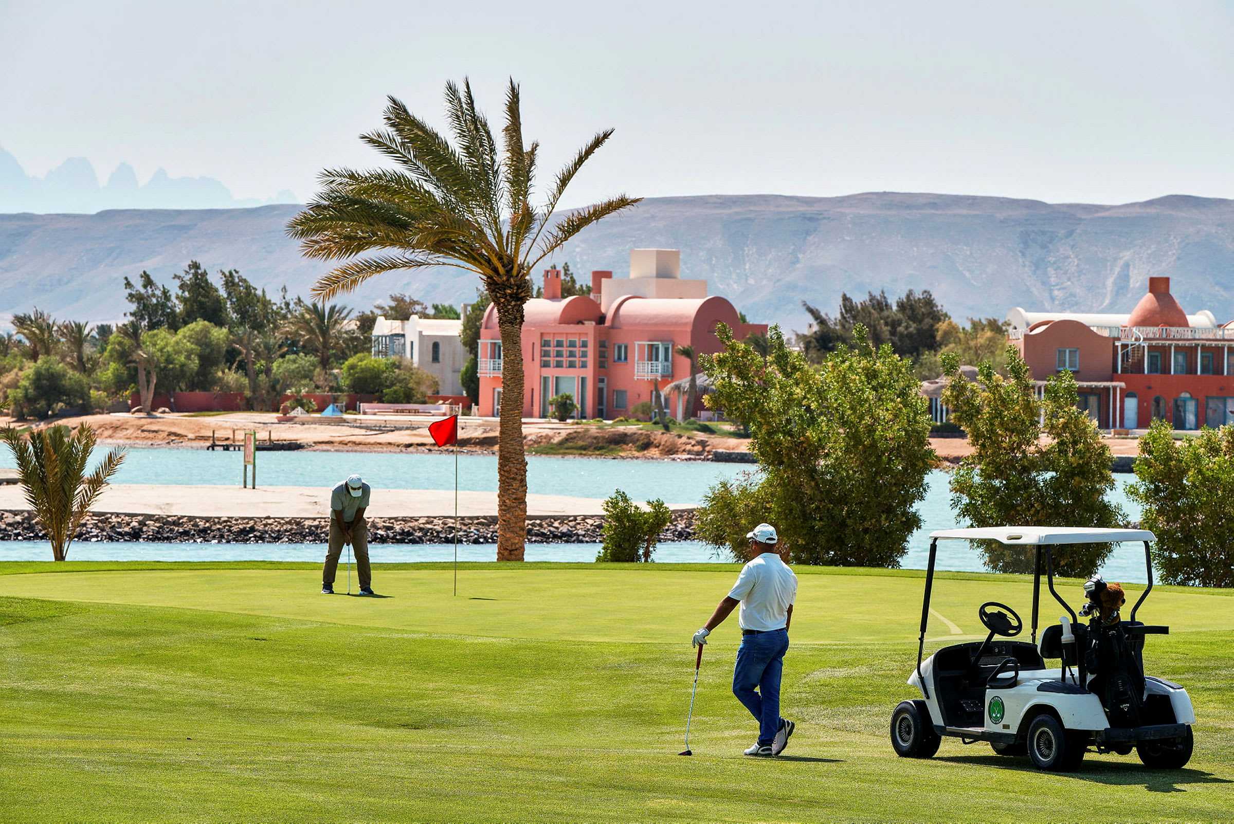 To personer spiller golf på en grøn golfbane nær vandet, med et paladsagtigt hus i baggrunden. (AI-genereret tekst)
