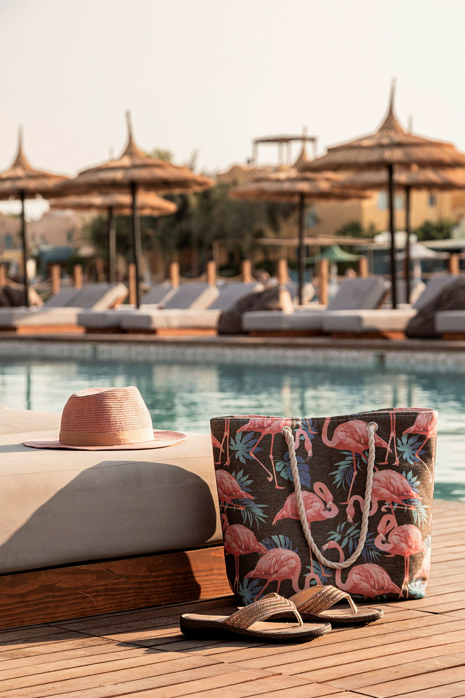 En hat, strandtaske og sandaler på en solstolsdyn ved en swimmingpool. (AI-genereret tekst)